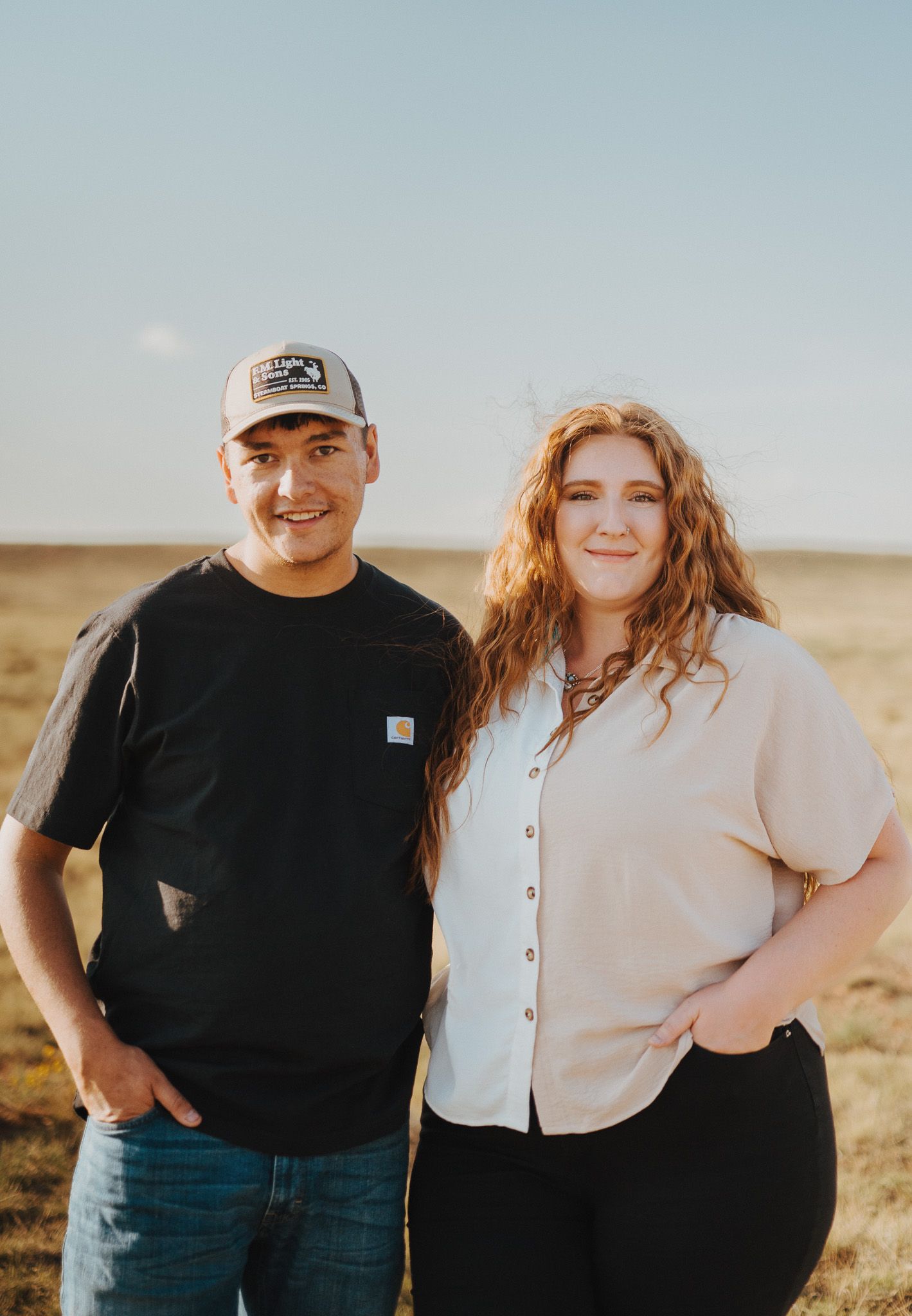 A couple poses together in a grassy landscape wearing contrasting black and beige outfits at sunset.