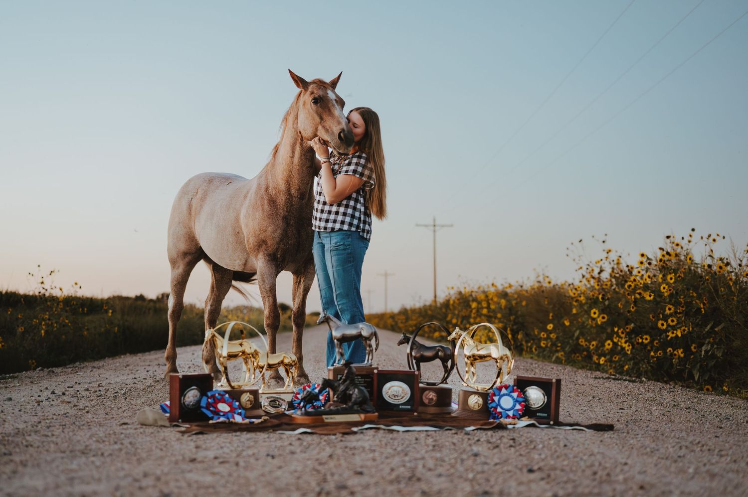 A young woman kissing a horse's nose in Lyman, Nebraska as they pose behind trophies and ribbons on the ground in front of them.