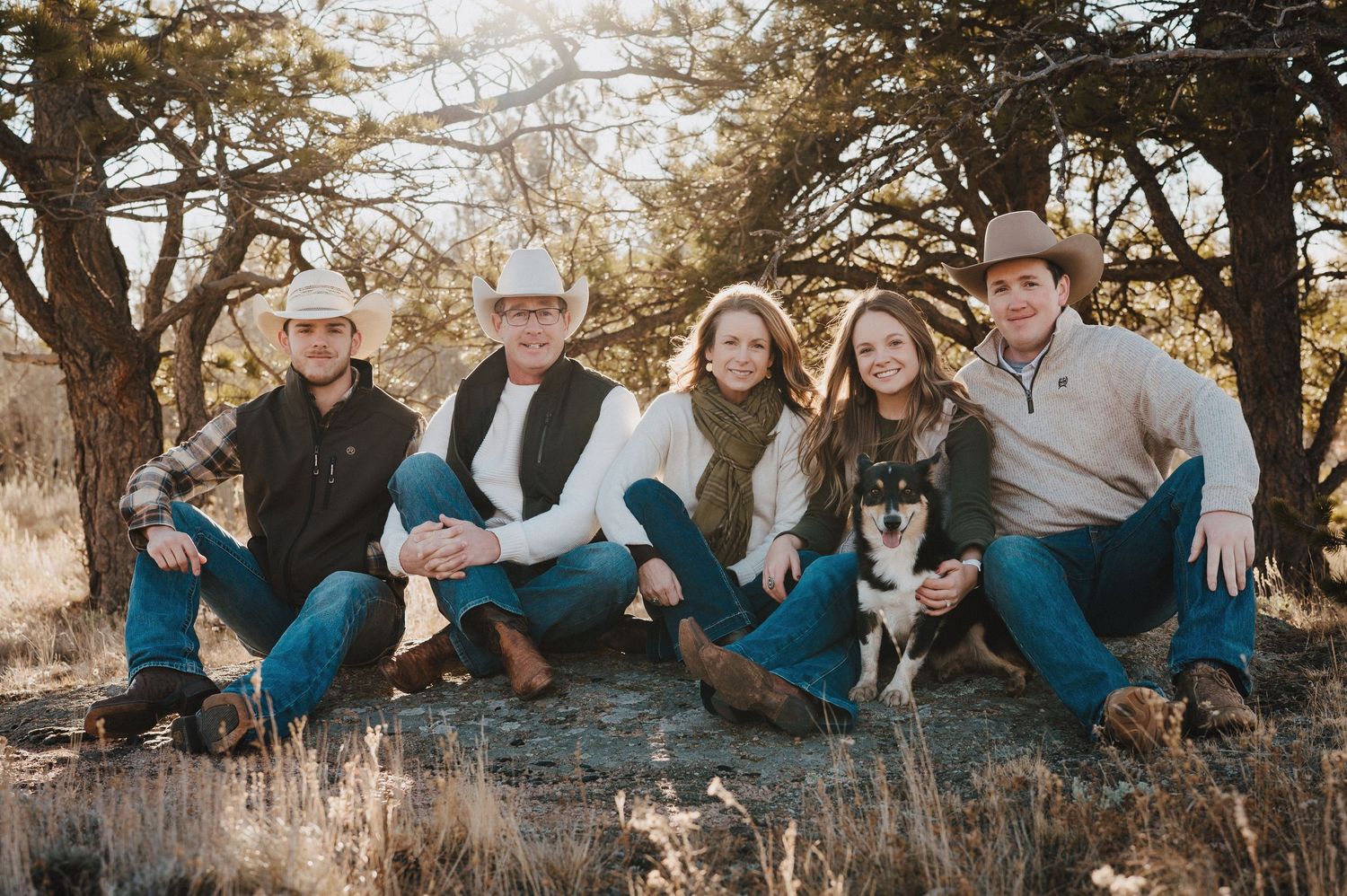 A group in coordinated blue jeans sits outdoors with their dog during golden hour in a natural setting.