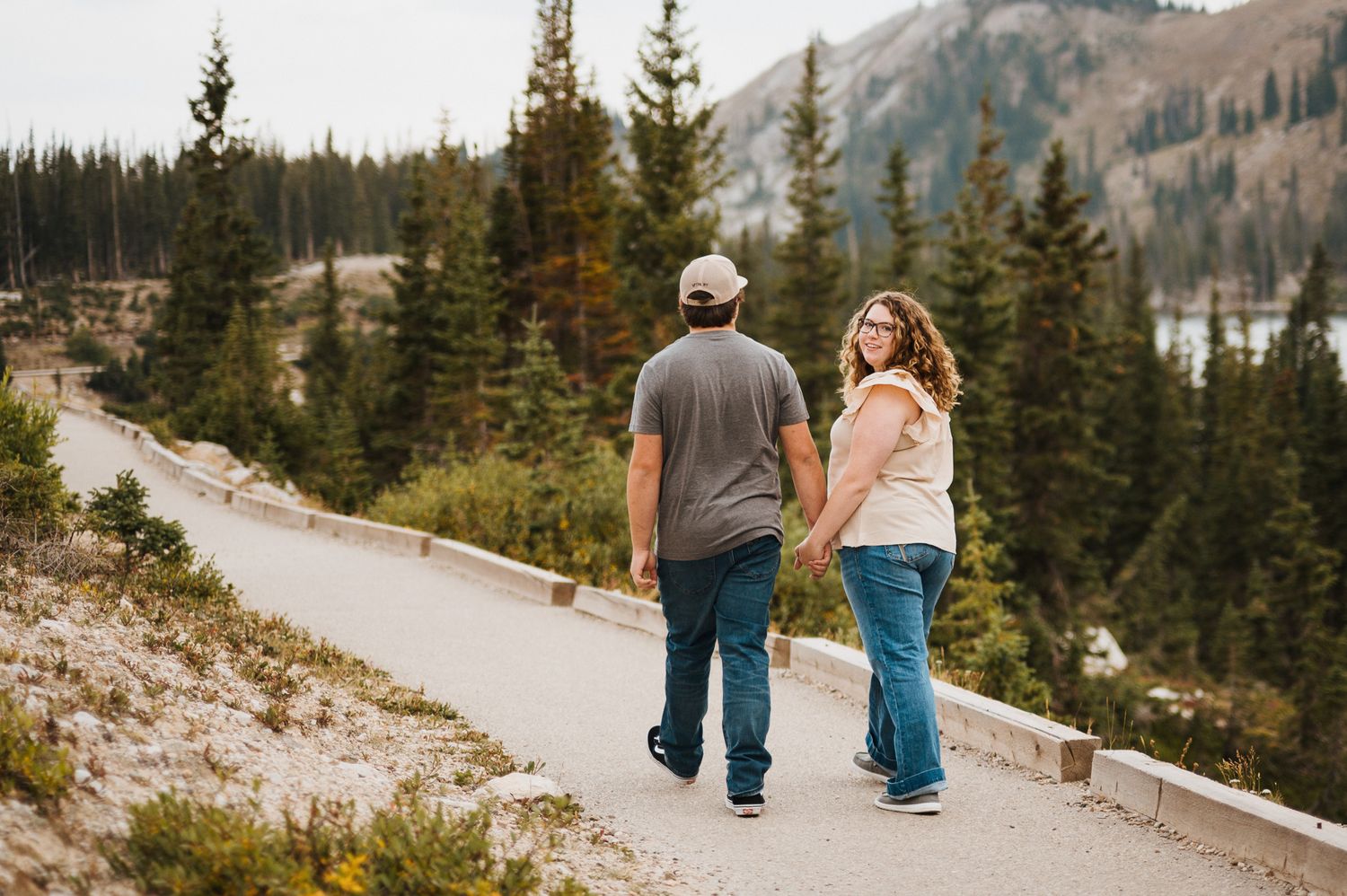 People walk along a scenic mountain trail surrounded by evergreen trees and rugged peaks in the distance.