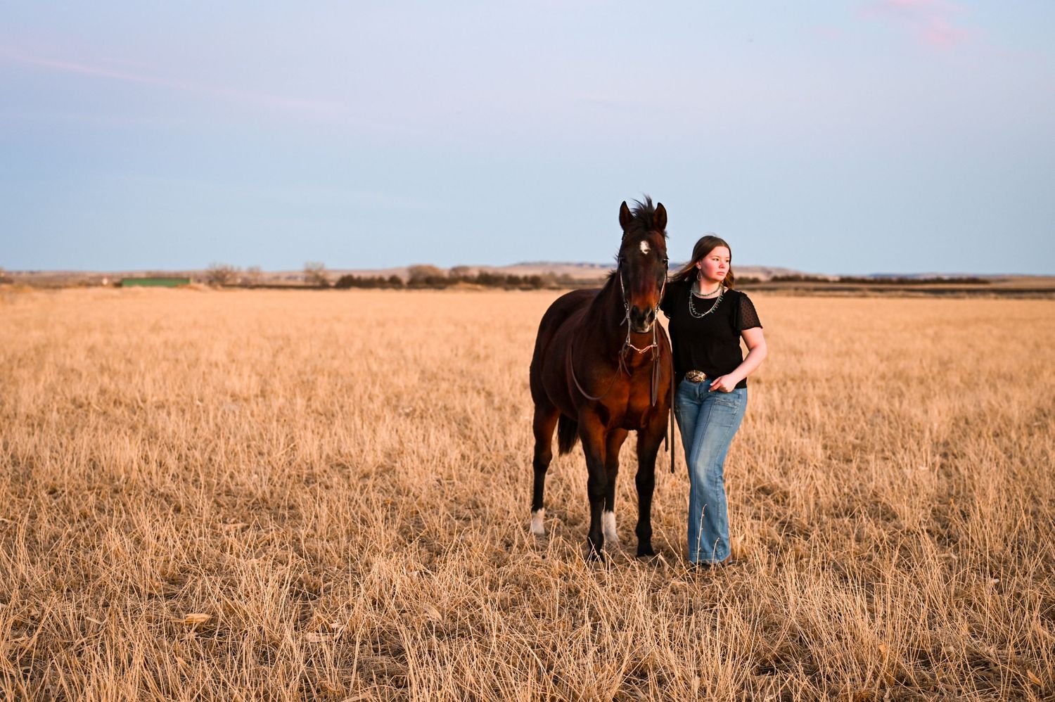 A woman standing next to a horse in a grassy field in Western Nebraska and looking into the distance.