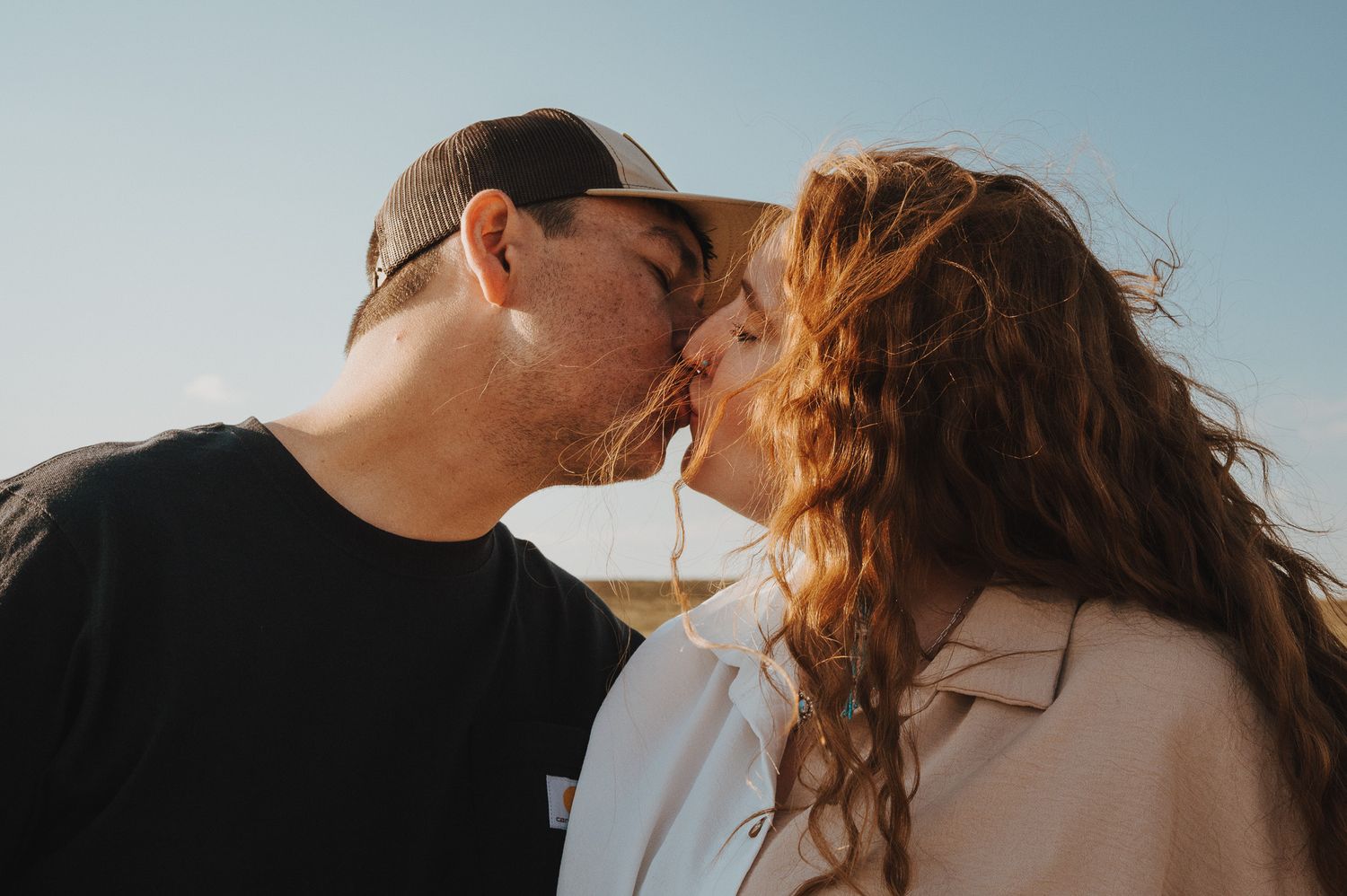A couple shares an intimate kiss against a soft blue sky background with sunlight creating a romantic silhouette.
