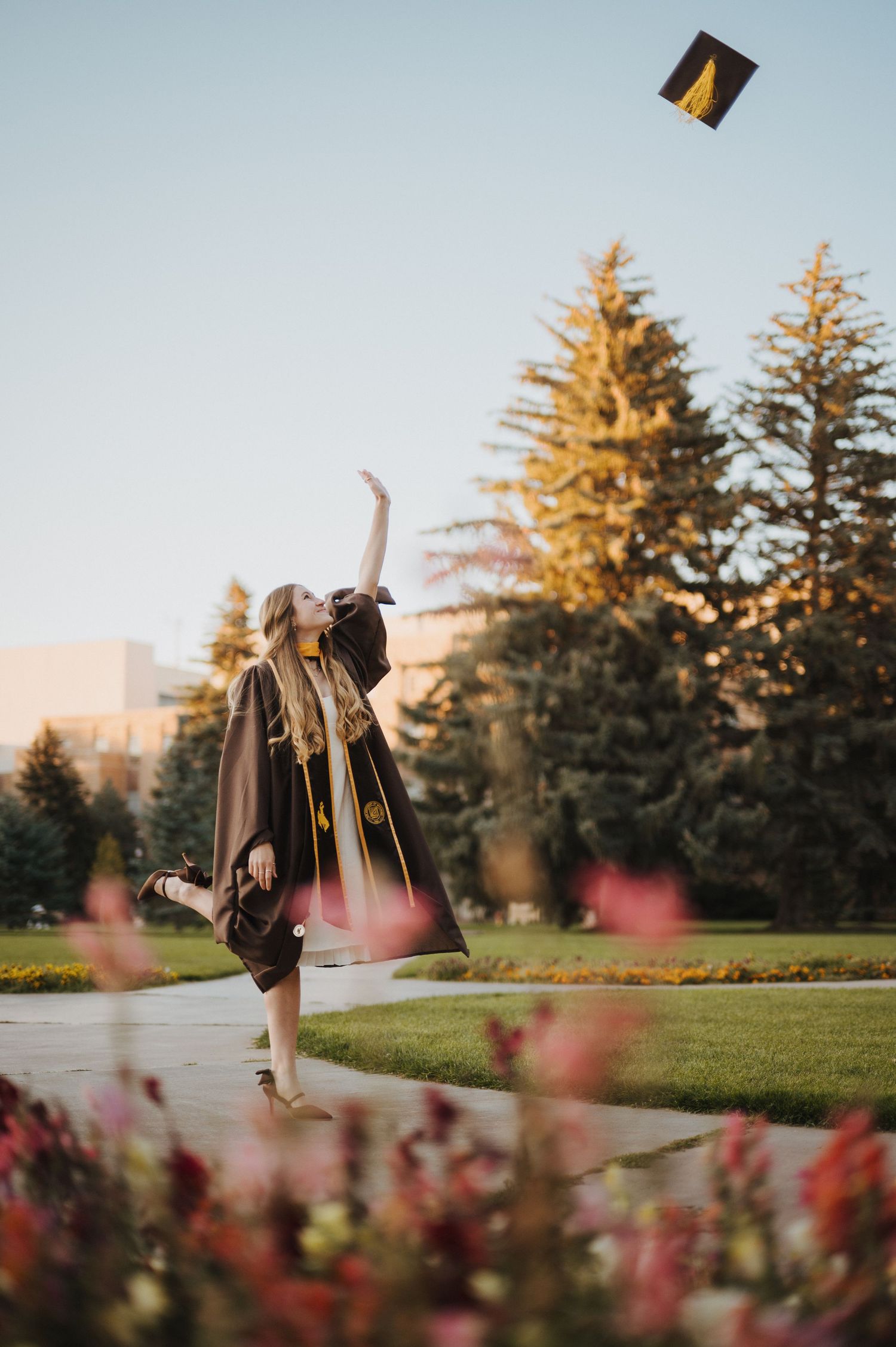 Graduate in cap and gown tosses graduation cap in golden sunlight with evergreen trees and pink flowers in foreground.
