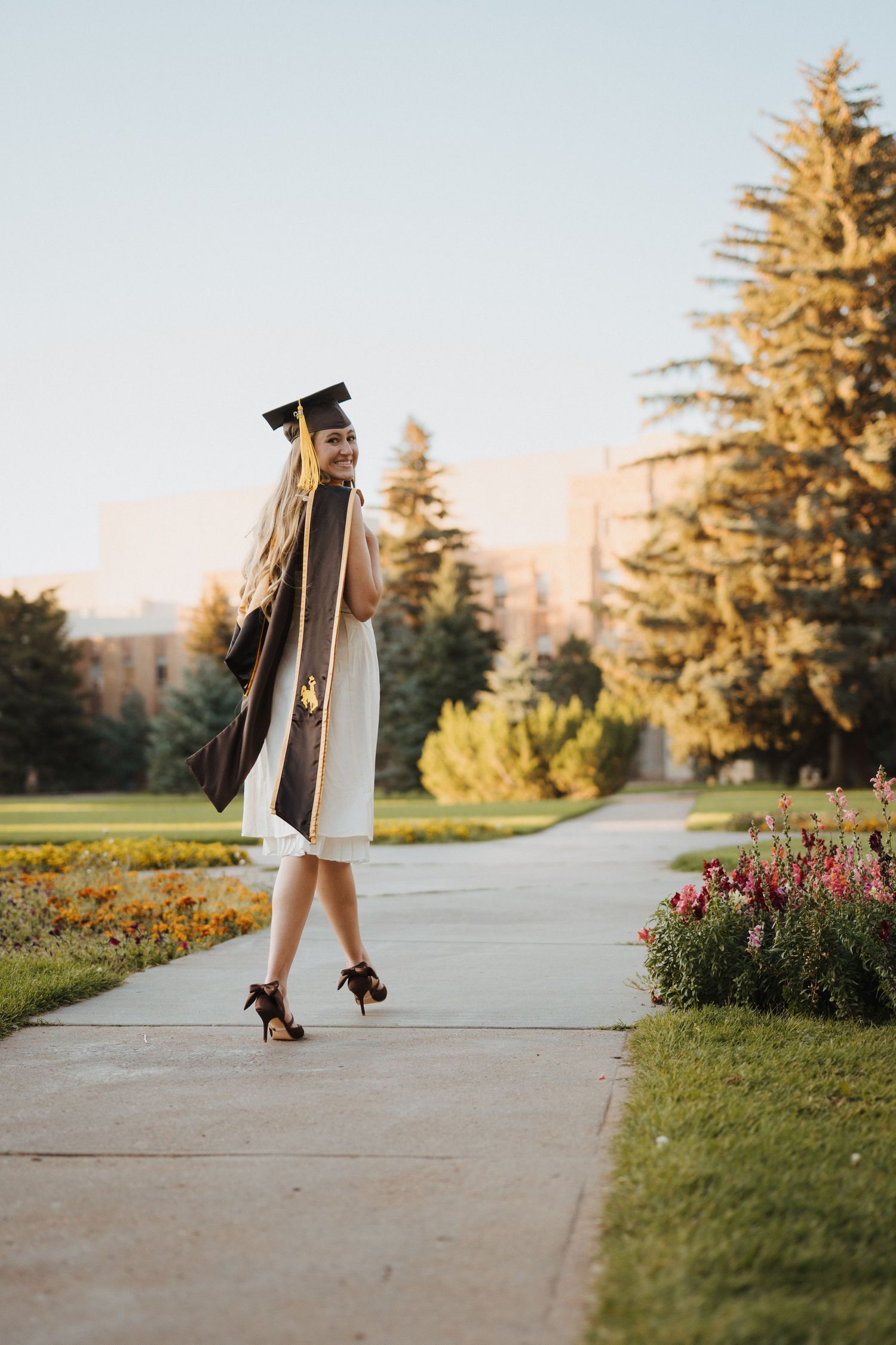 Graduate in cap and gown twirls on a garden path during golden hour with evergreen trees in background.