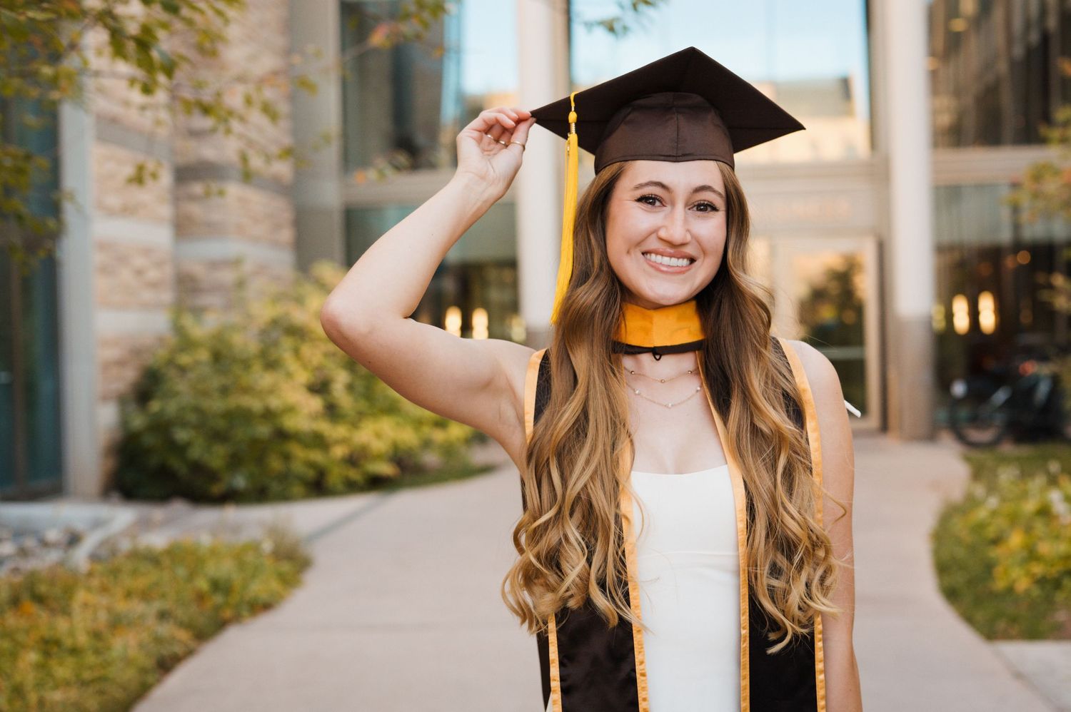 A graduate in a black cap and gown stands smiling outside a stone building during a sunny graduation day.