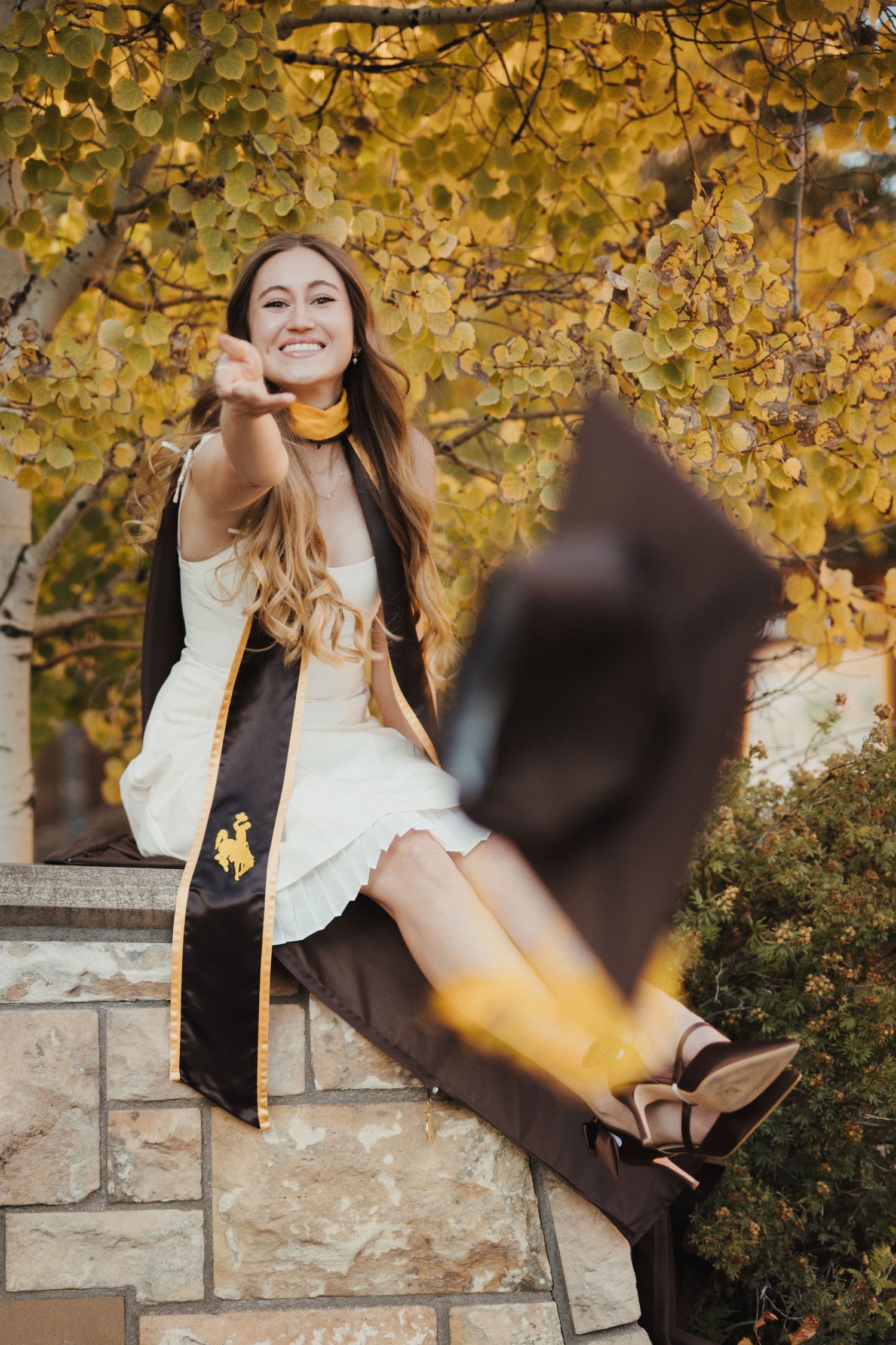 A woman sitting on a stone ledge, throwing her graduation cap toward the camera.