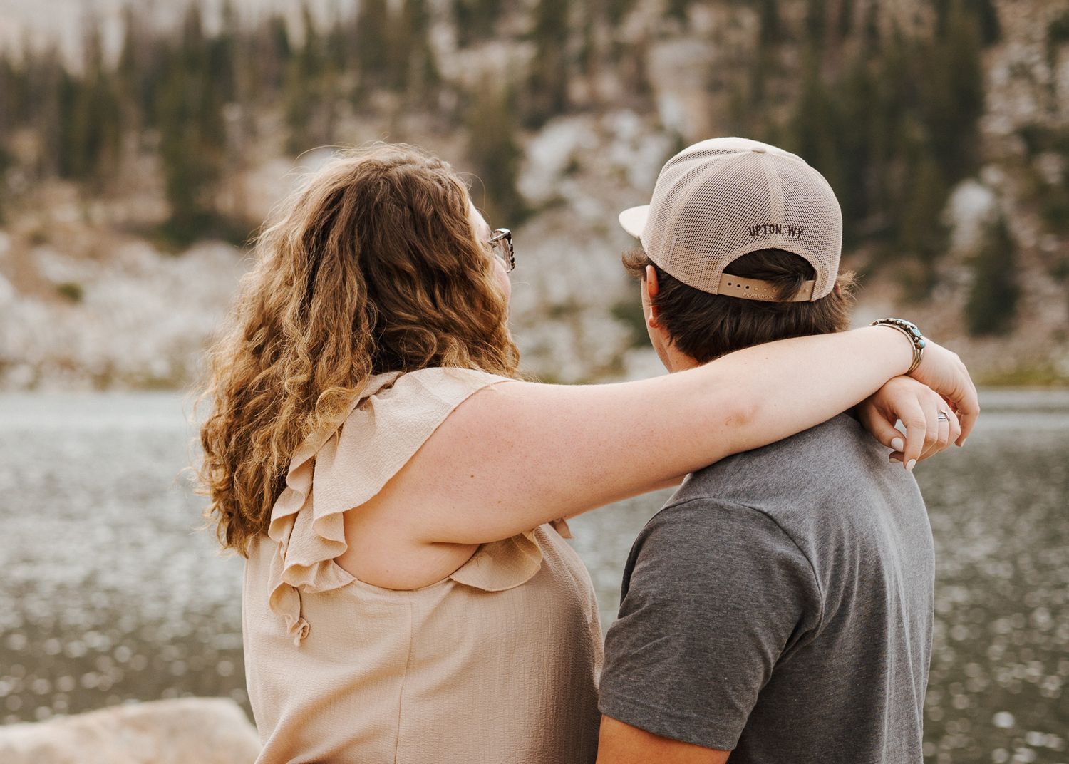 A couple stands together in an outdoor mountain setting with evergreen trees and snow visible in the background.