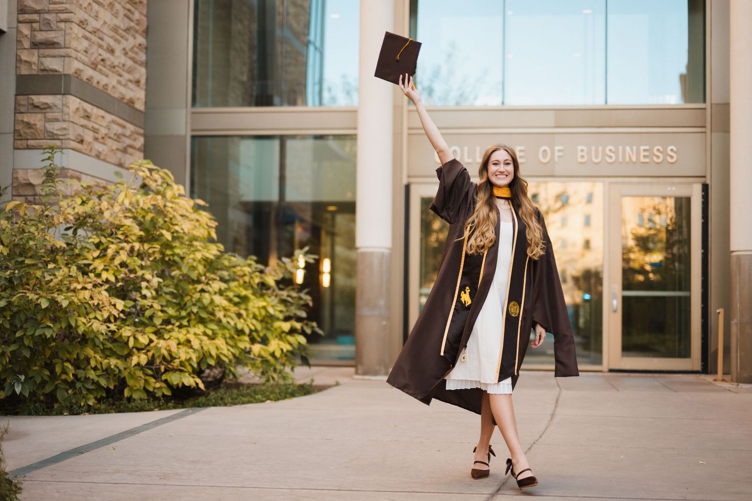 A woman in graduation regalia smiles and holds up her cap as she walks down a path.