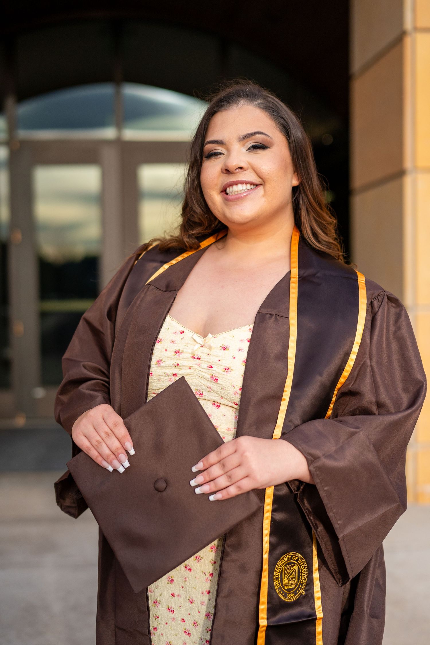 Graduate in brown academic regalia and polka dot dress poses for celebratory graduation photos outdoors.
