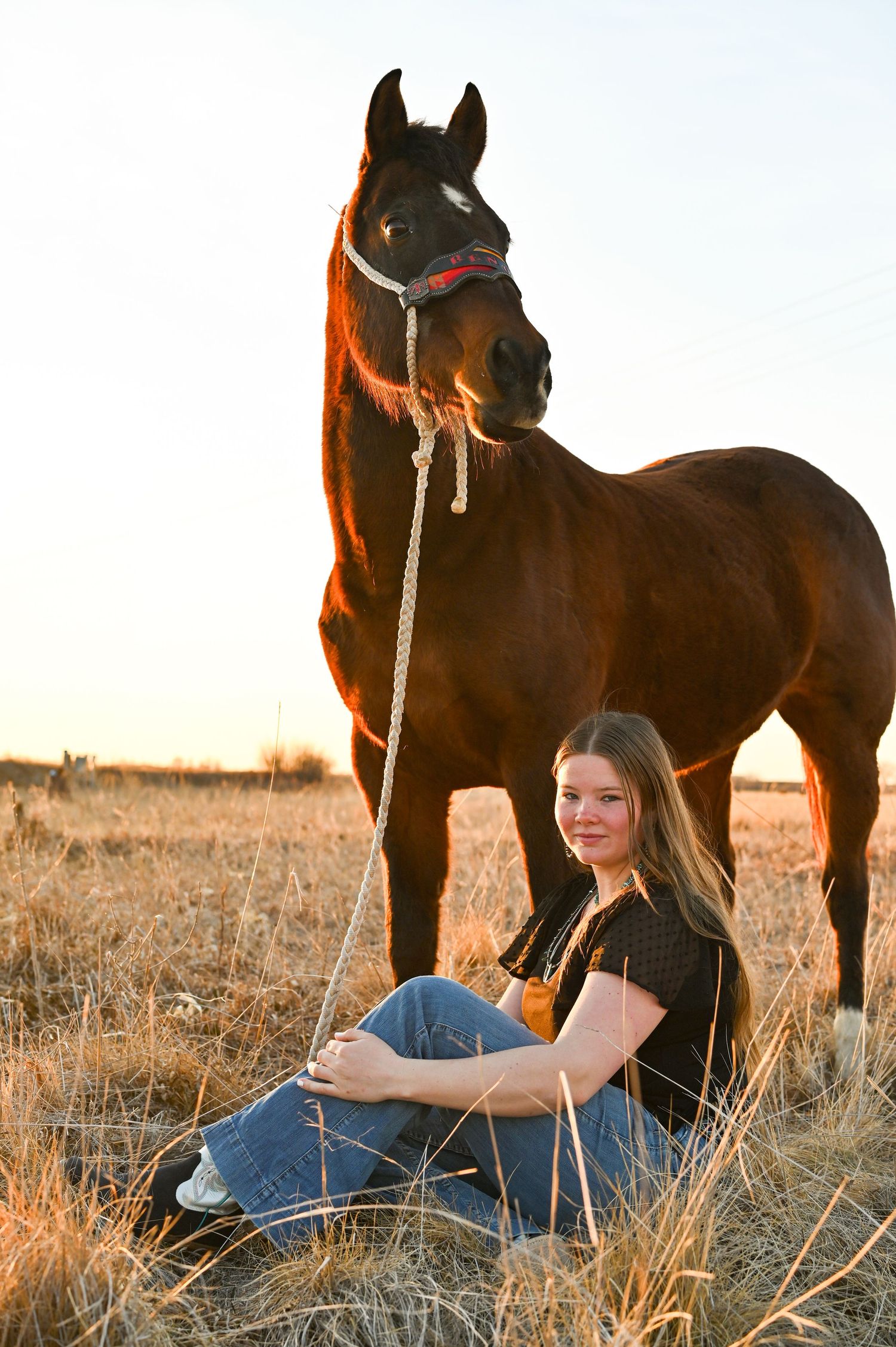 A woman in casual attire sits in a field in Western Nebraska at sunset with a dark horse standing behind her.