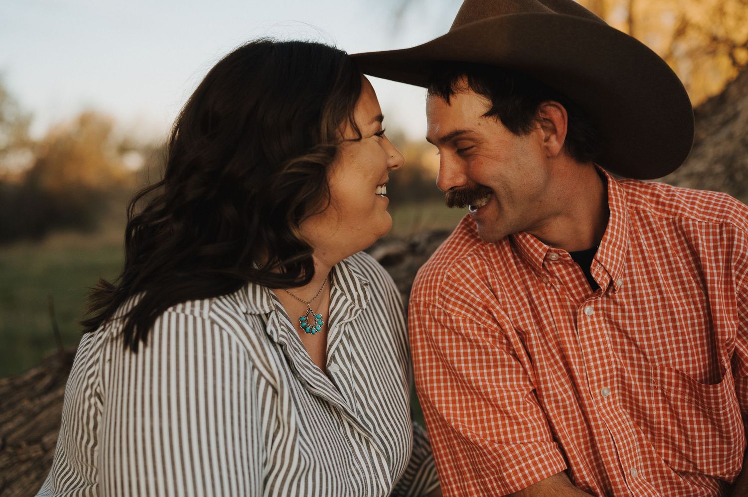 Couple in western attire share romantic moments during golden hour sunset photo session in rustic outdoor setting.