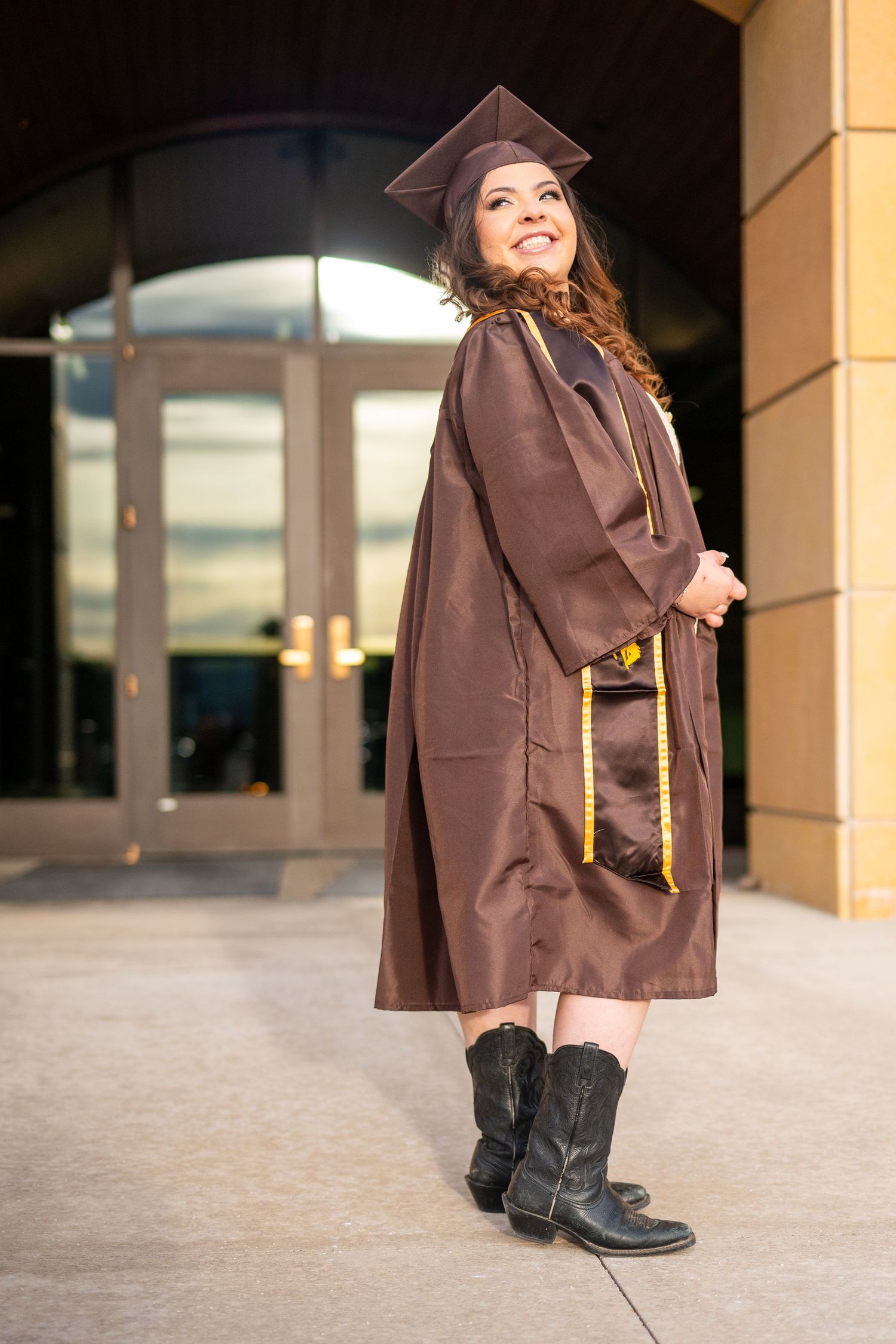 A woman in a brown gown and black boots poses against a beige wall and columns in Laramie, Wyoming.