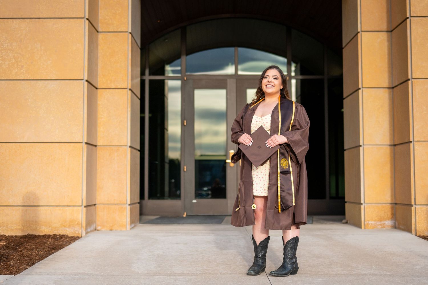 A college senior woman wearing graduation regalia smiles while posing in front of the University of Wyoming College of Law.