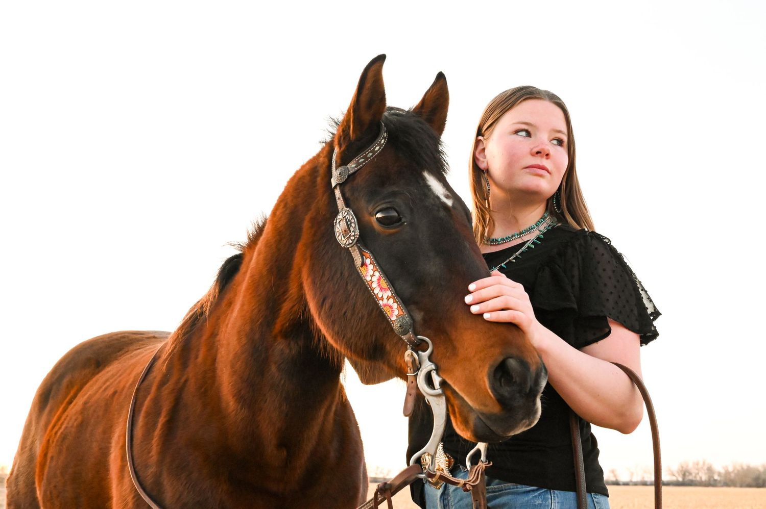 A woman and her dark horse share a tender moment during sunset in a rustic outdoor setting.