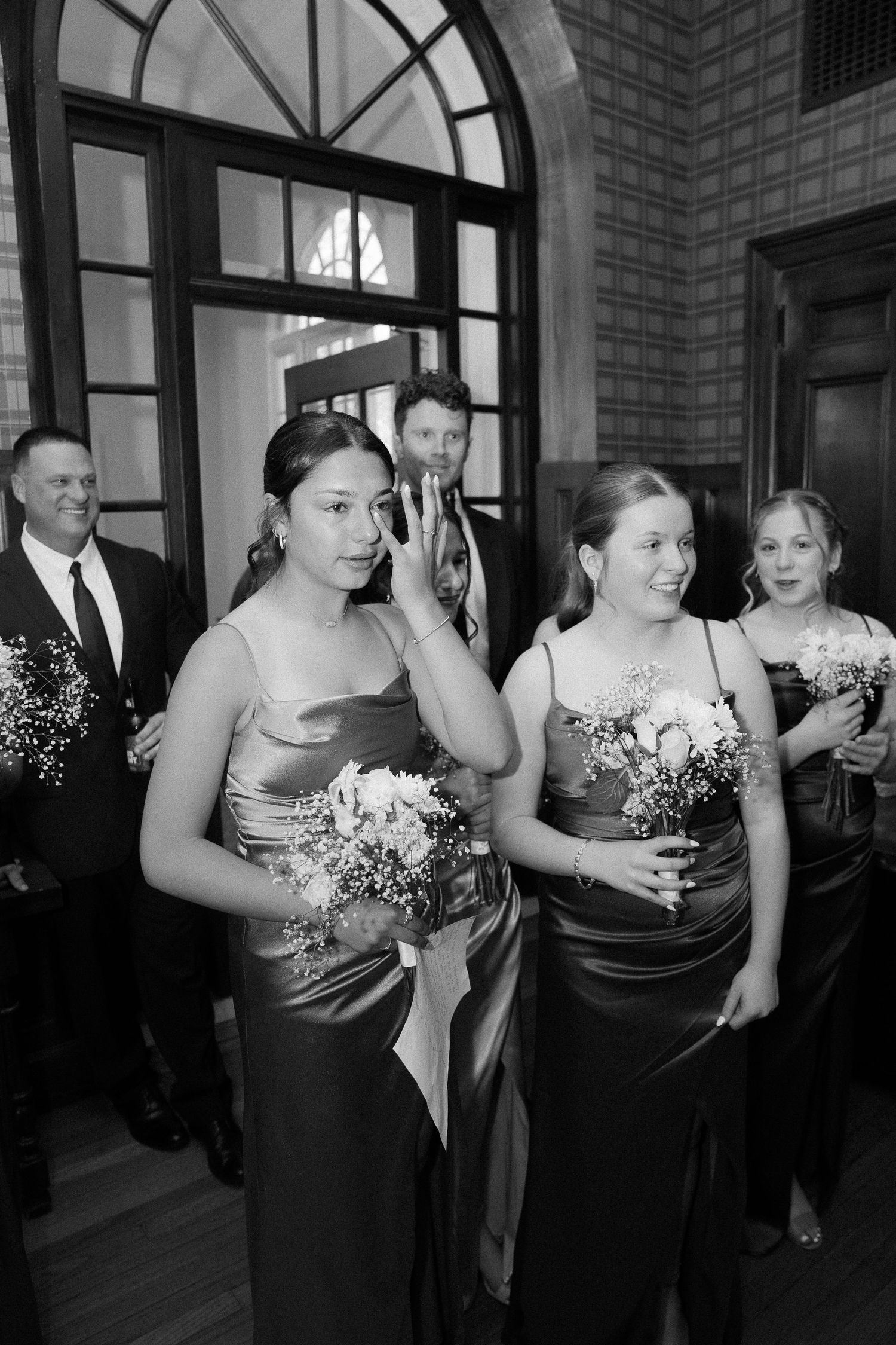 Bridesmaids in elegant satin gowns holding floral bouquets walk in procession at a wedding ceremony in black and white.