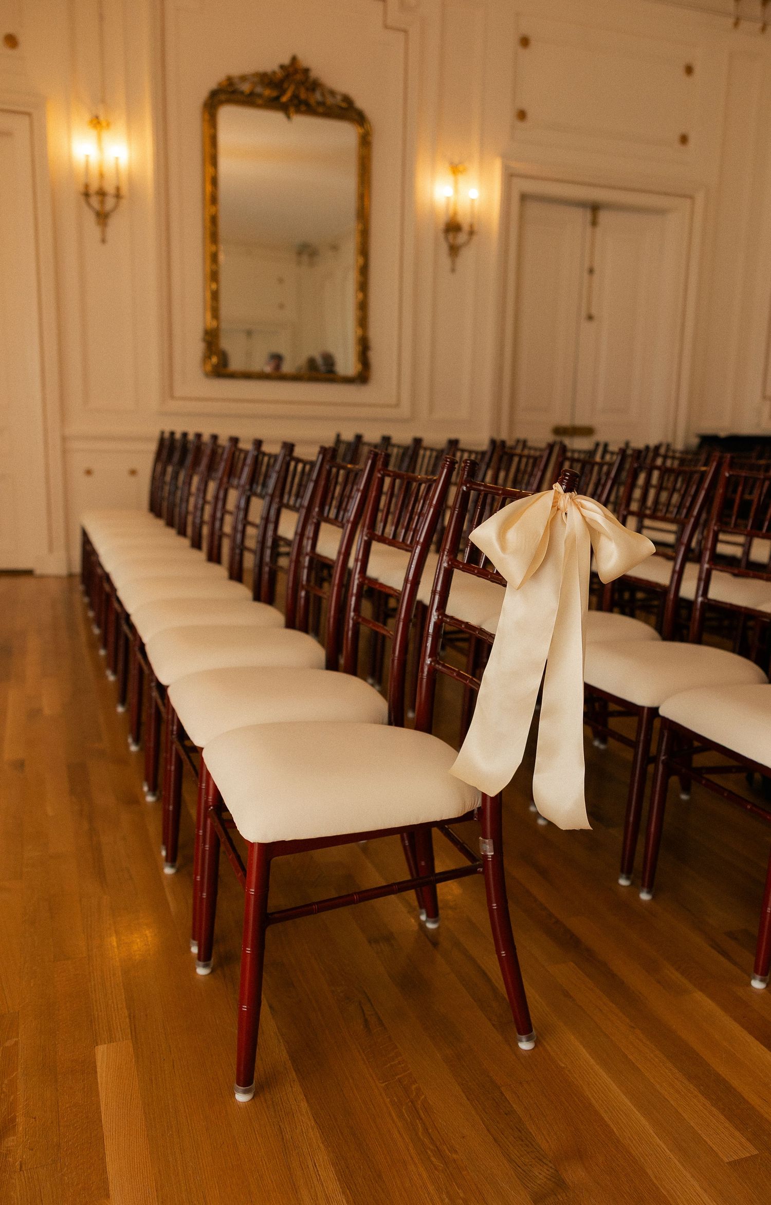 Elegant wedding ceremony chairs with ivory cushions and satin bow, arranged in rows in a grand ballroom with gold mirrors.