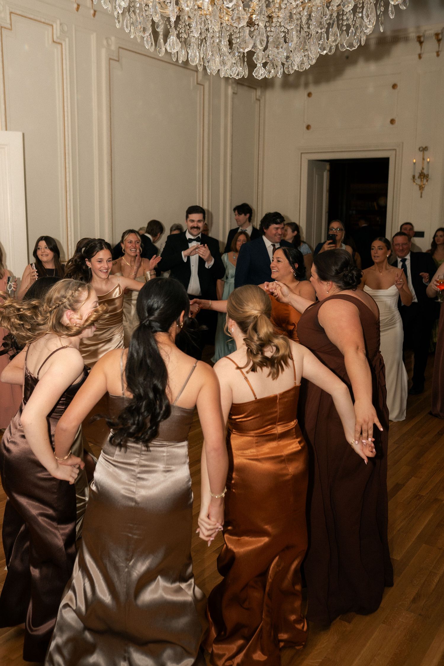 Bridesmaids in satin gowns dance joyfully at an elegant wedding reception under a crystal chandelier.