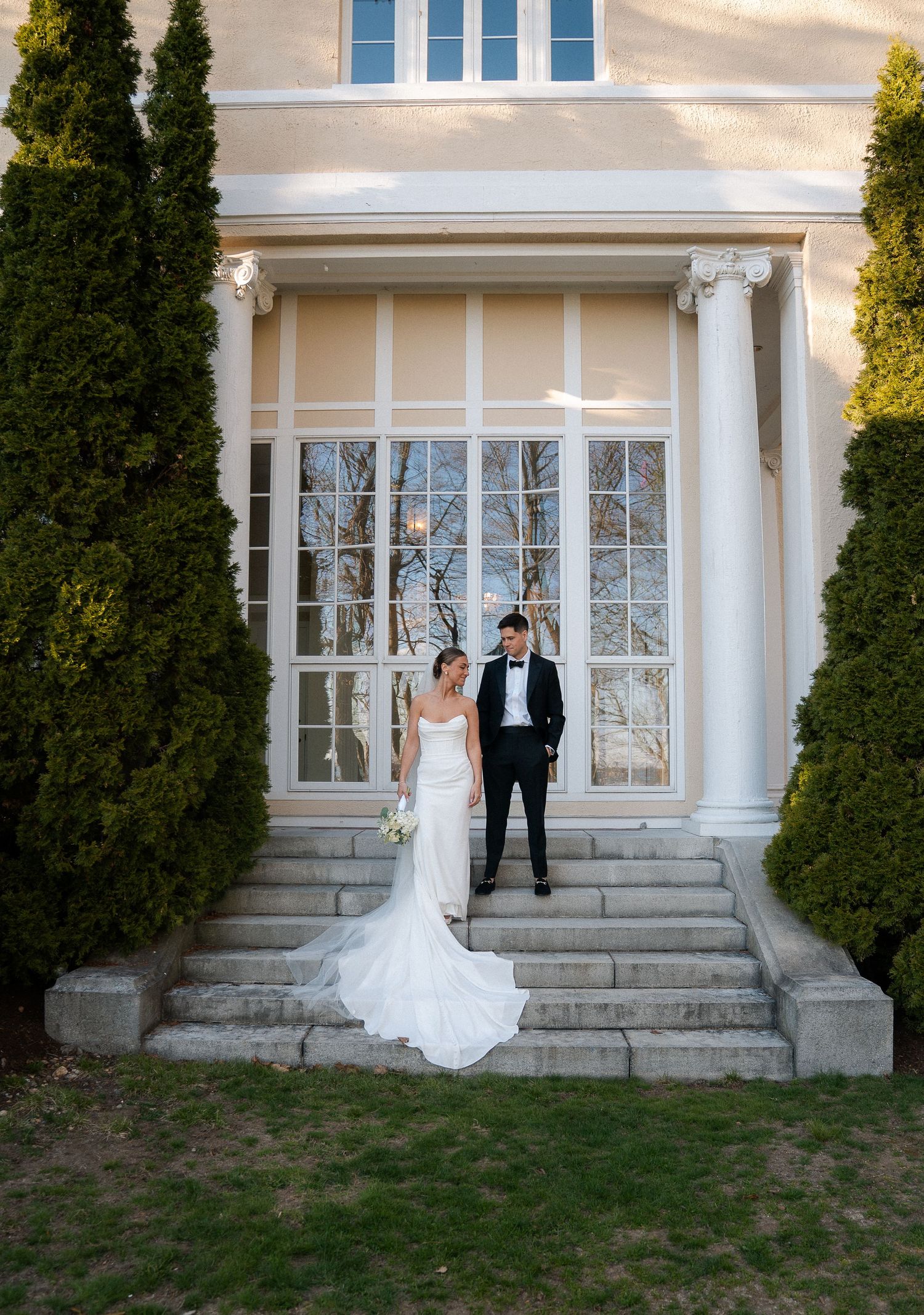 Bride and groom pose on stone steps of elegant venue flanked by tall cypress trees on their wedding day.