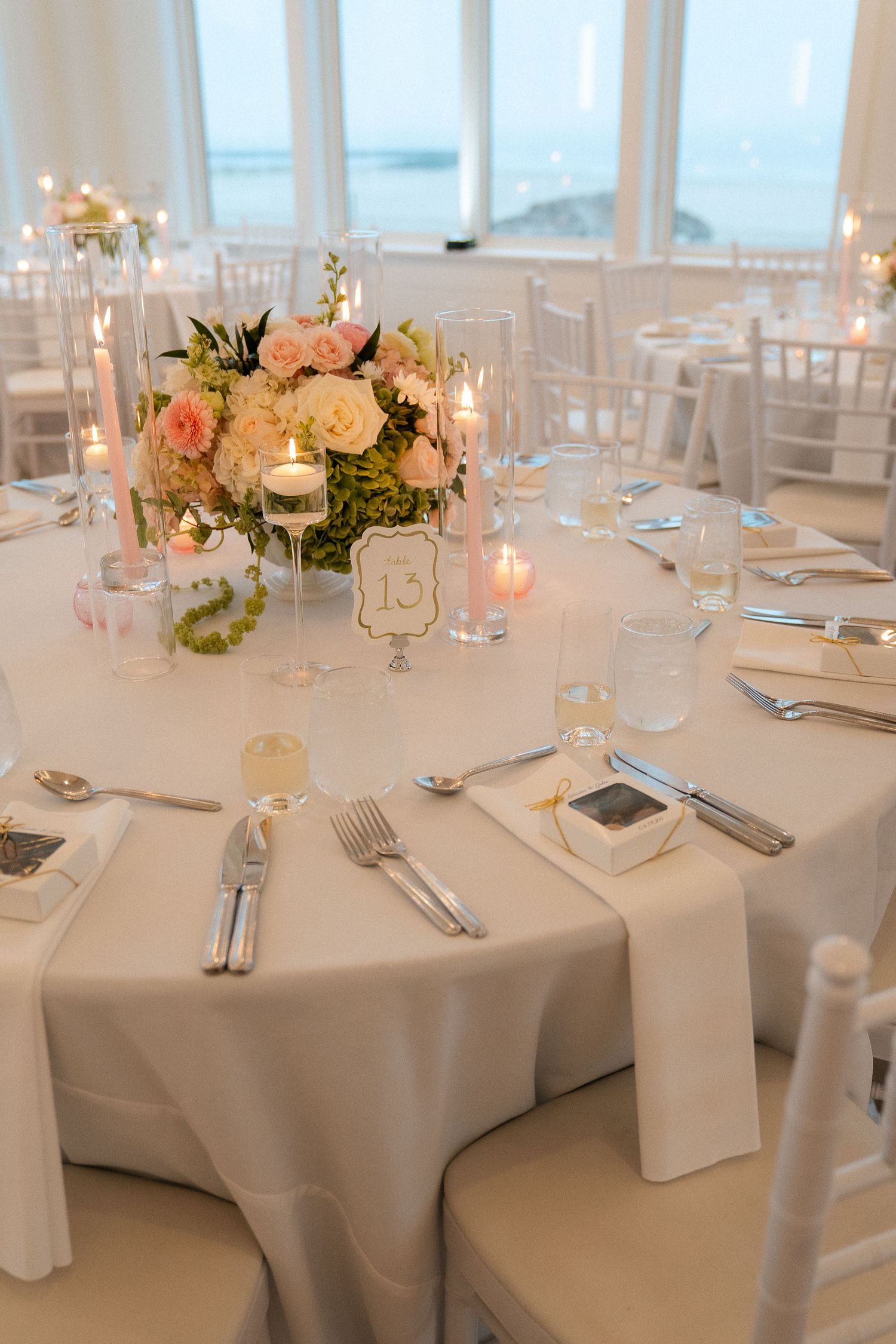 Elegant wedding reception table set with white linens, pink floral centerpiece, candles, and table number 13 by oceanview windows.