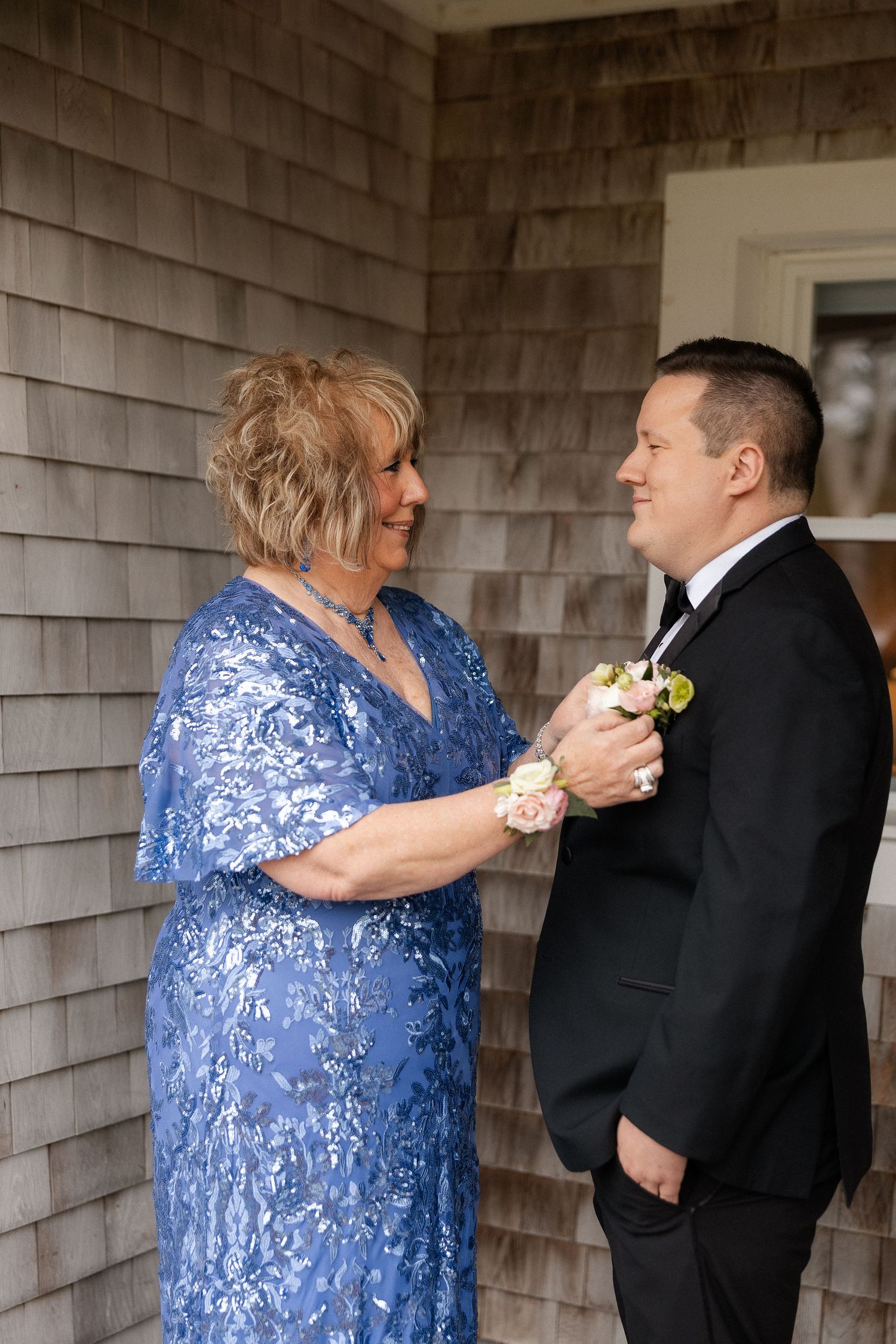 Mother in blue lace dress pinning boutonniere on groom in black tuxedo outside shingle-sided building.