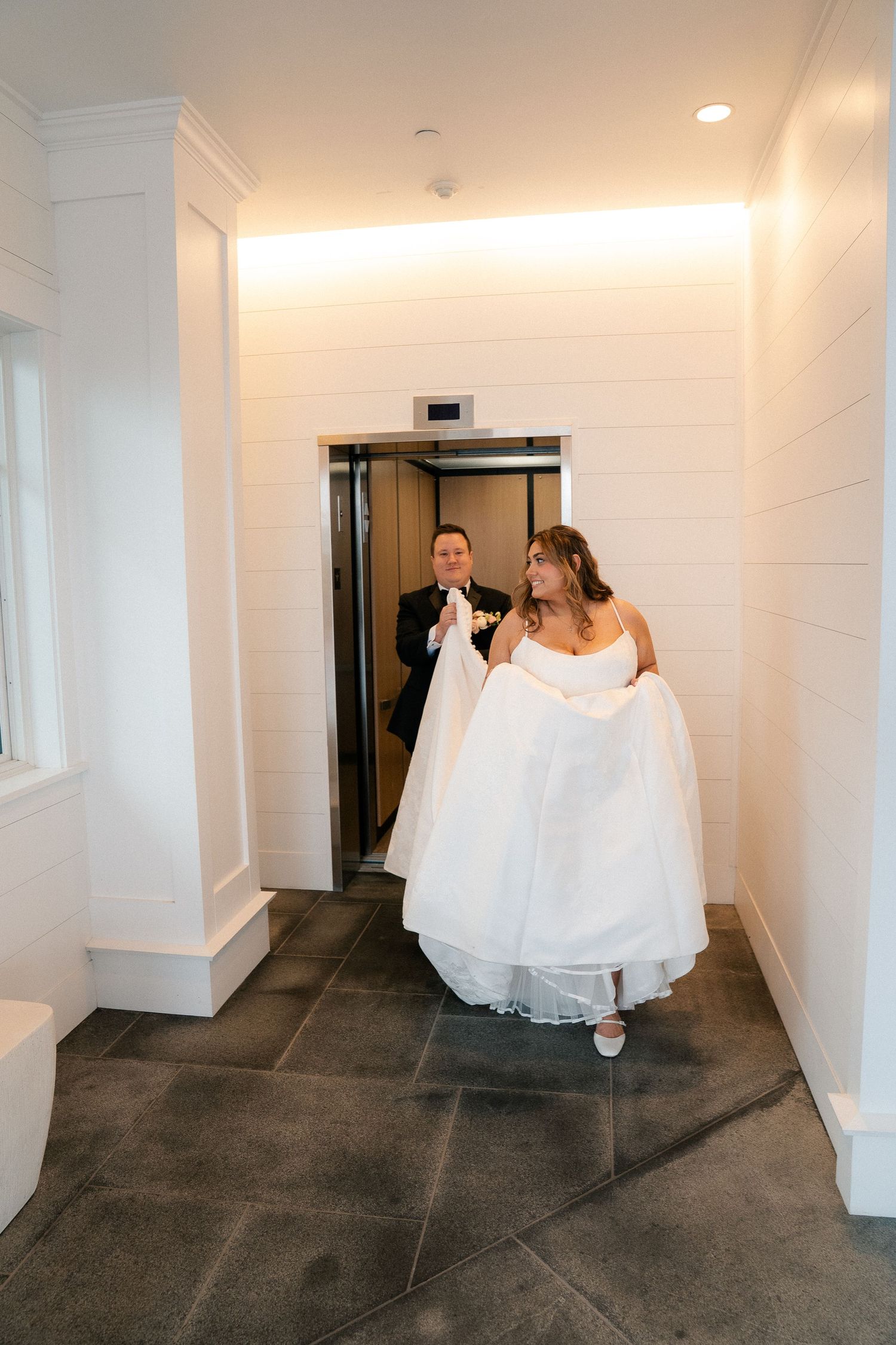 Bride lifting wedding gown exits elevator with groom in elegant white venue hallway.