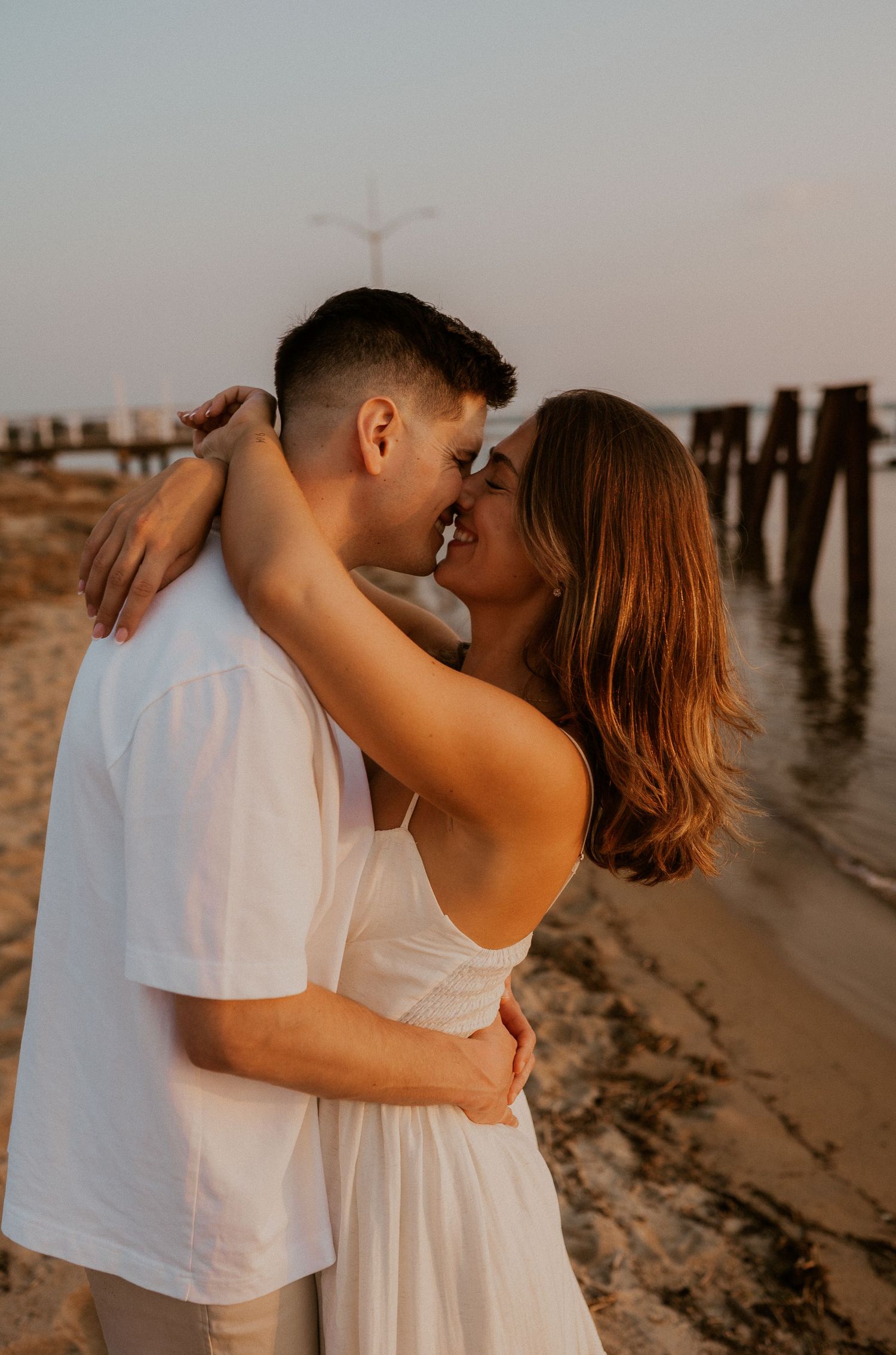 A romantic couple embraces on a beach at sunset near wooden pier posts.