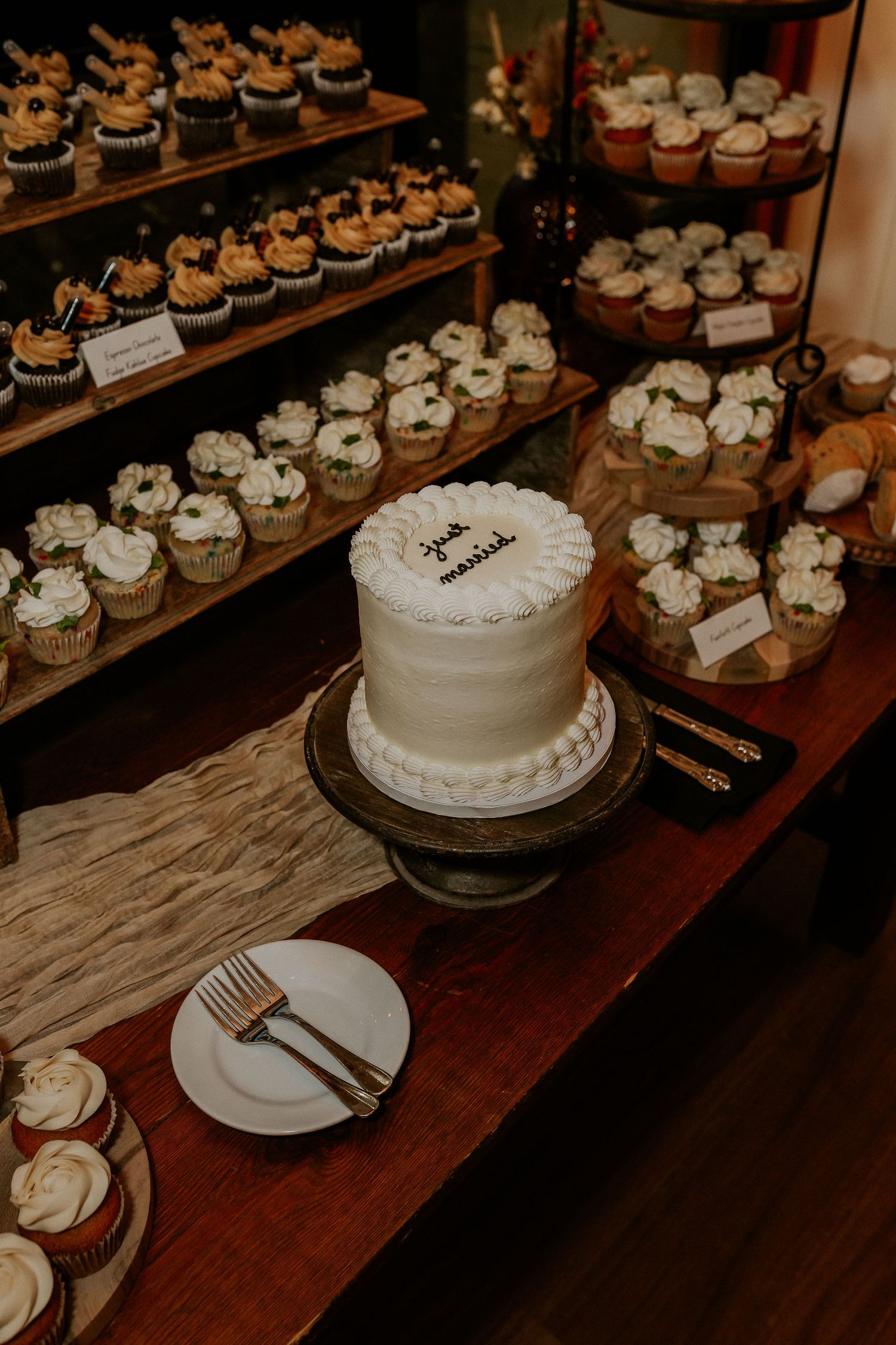 Rustic wooden table displaying an elegant white wedding cake and matching cupcakes arranged on multiple tiers.