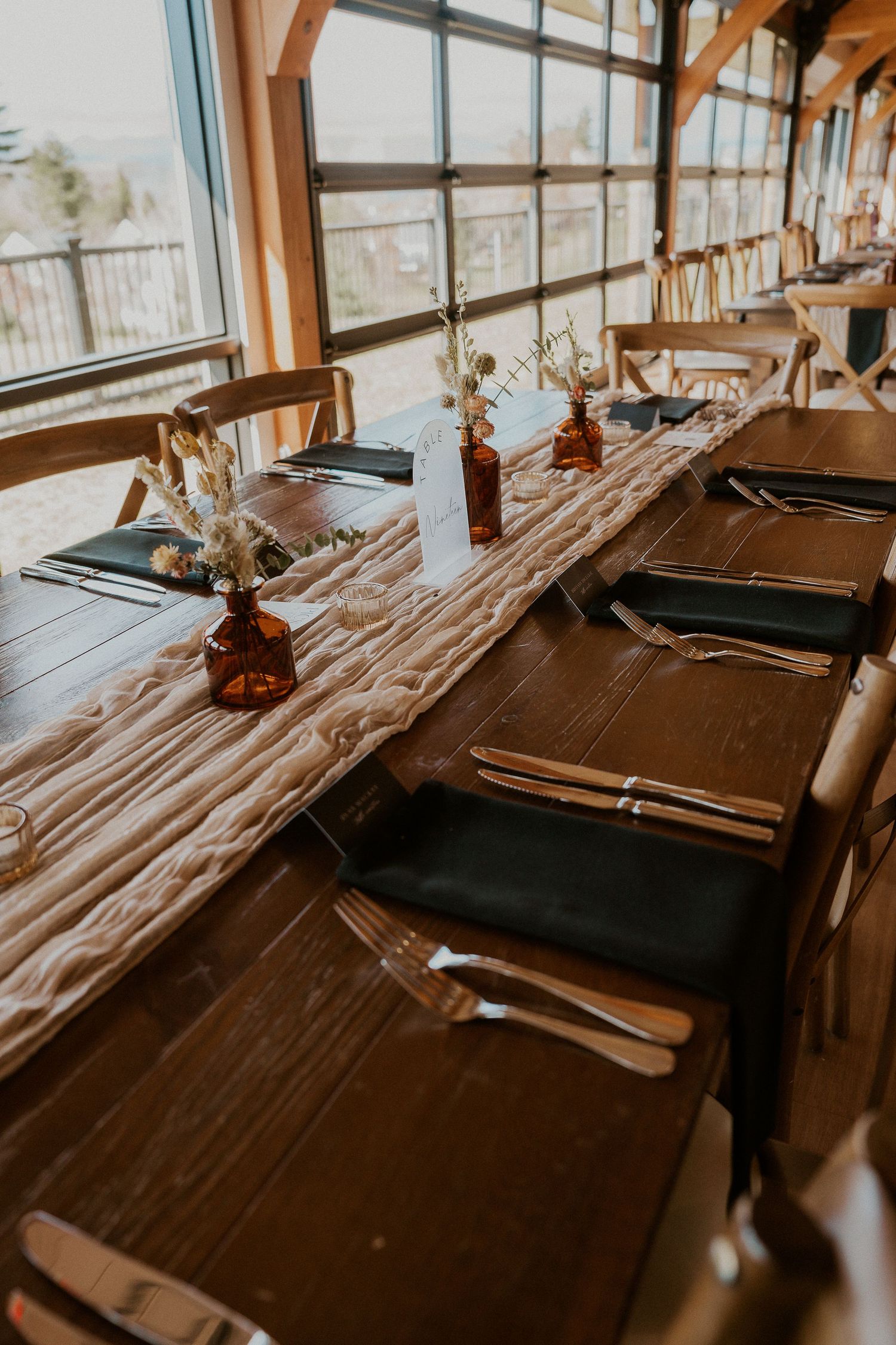 Rustic wooden table setup with dark place settings and window lighting in wedding venue.