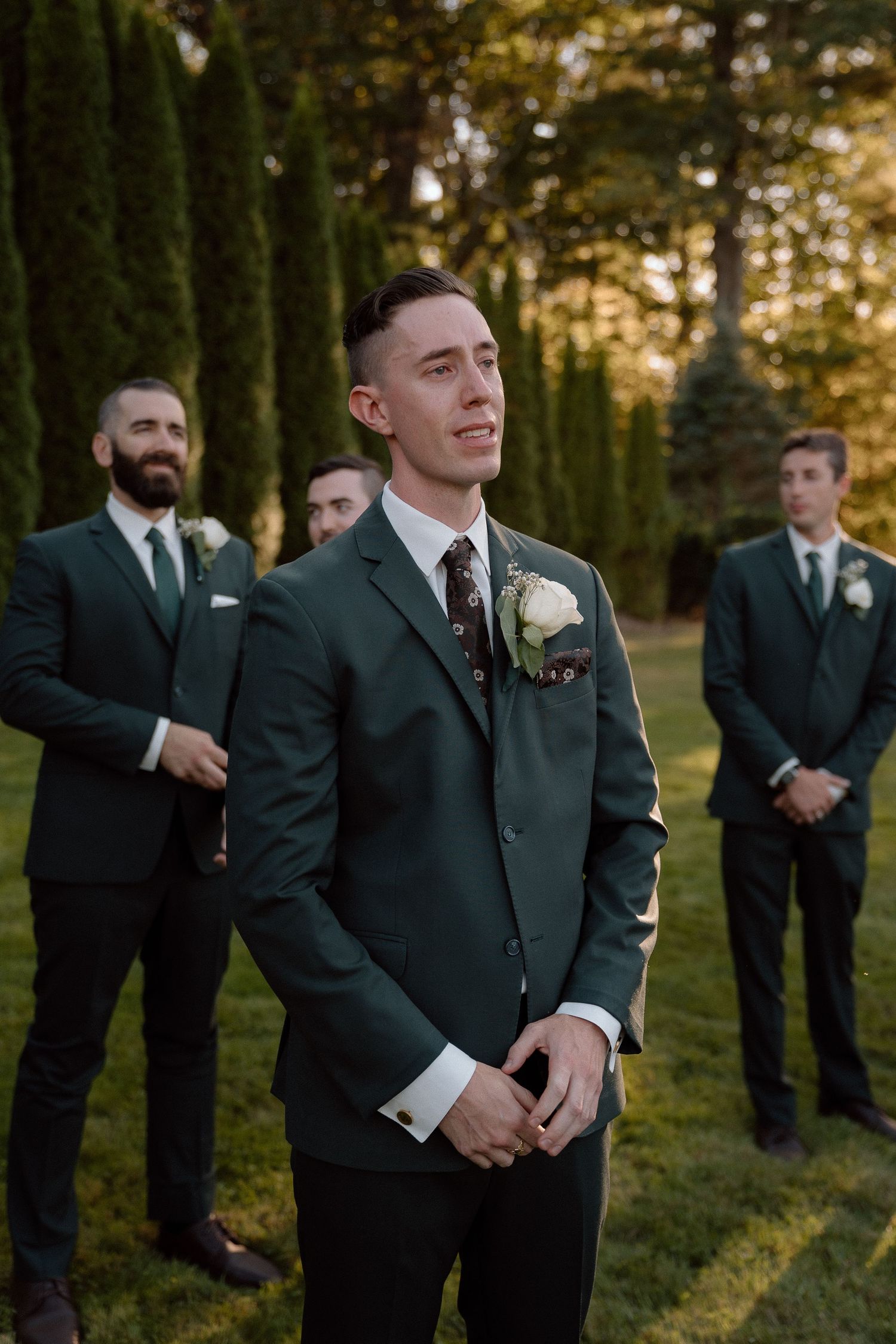 Groomsmen in dark suits stand in formation on a lawn during golden hour.