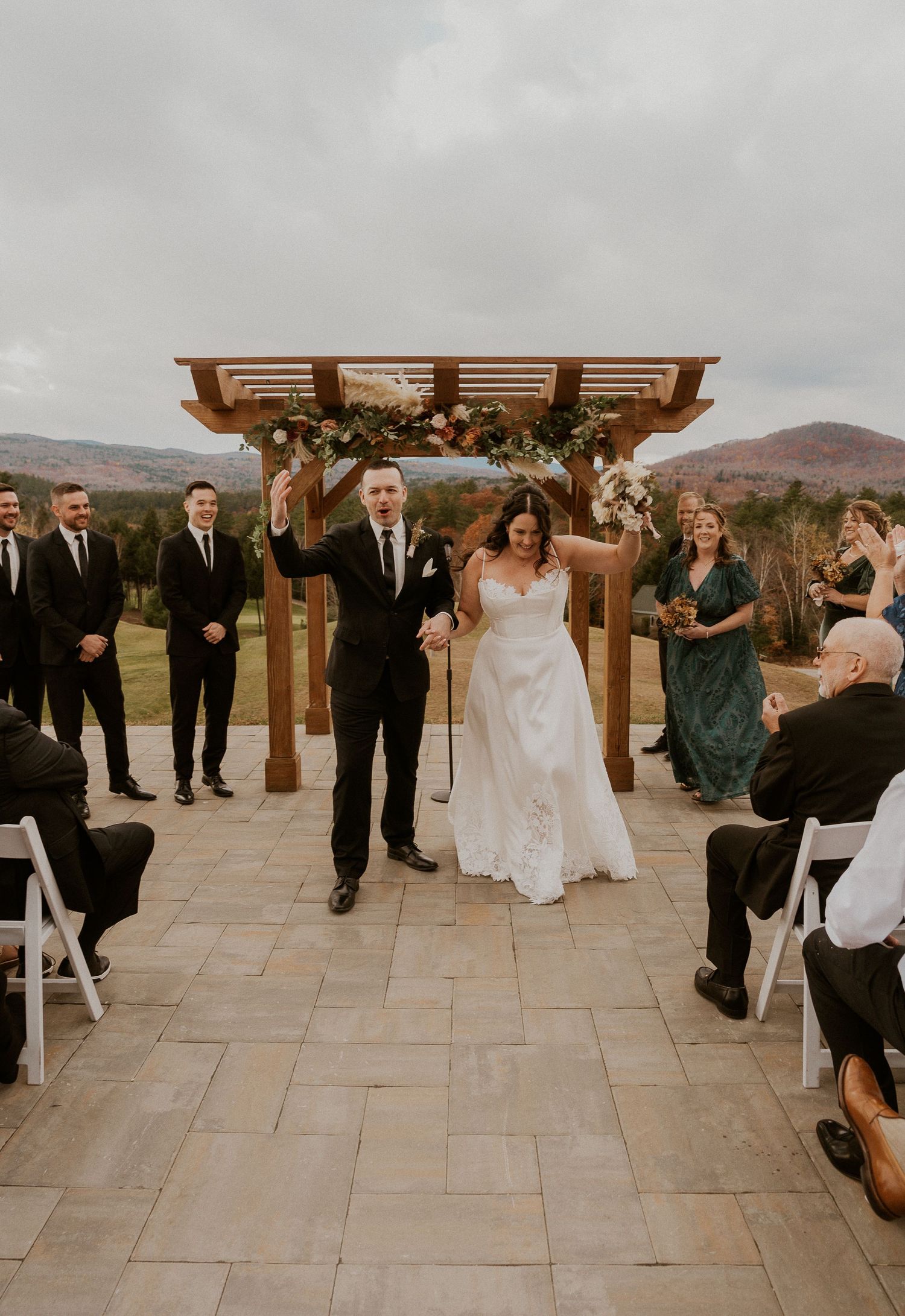 Wedding ceremony guests seated outdoors under wooden pergola with mountain views in the background.