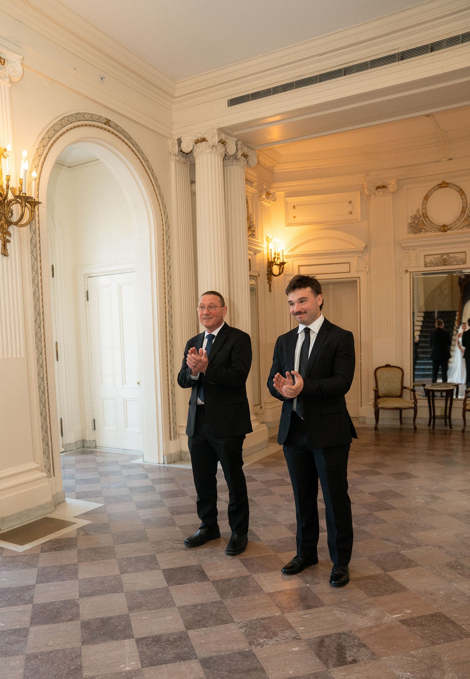Two men in black suits clapping in an elegant white ballroom with arched doorways and ornate decor.