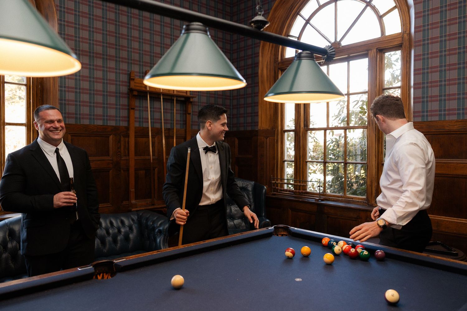 Three men in formal attire laughing and playing snooker in an elegant wood-paneled billiards room.