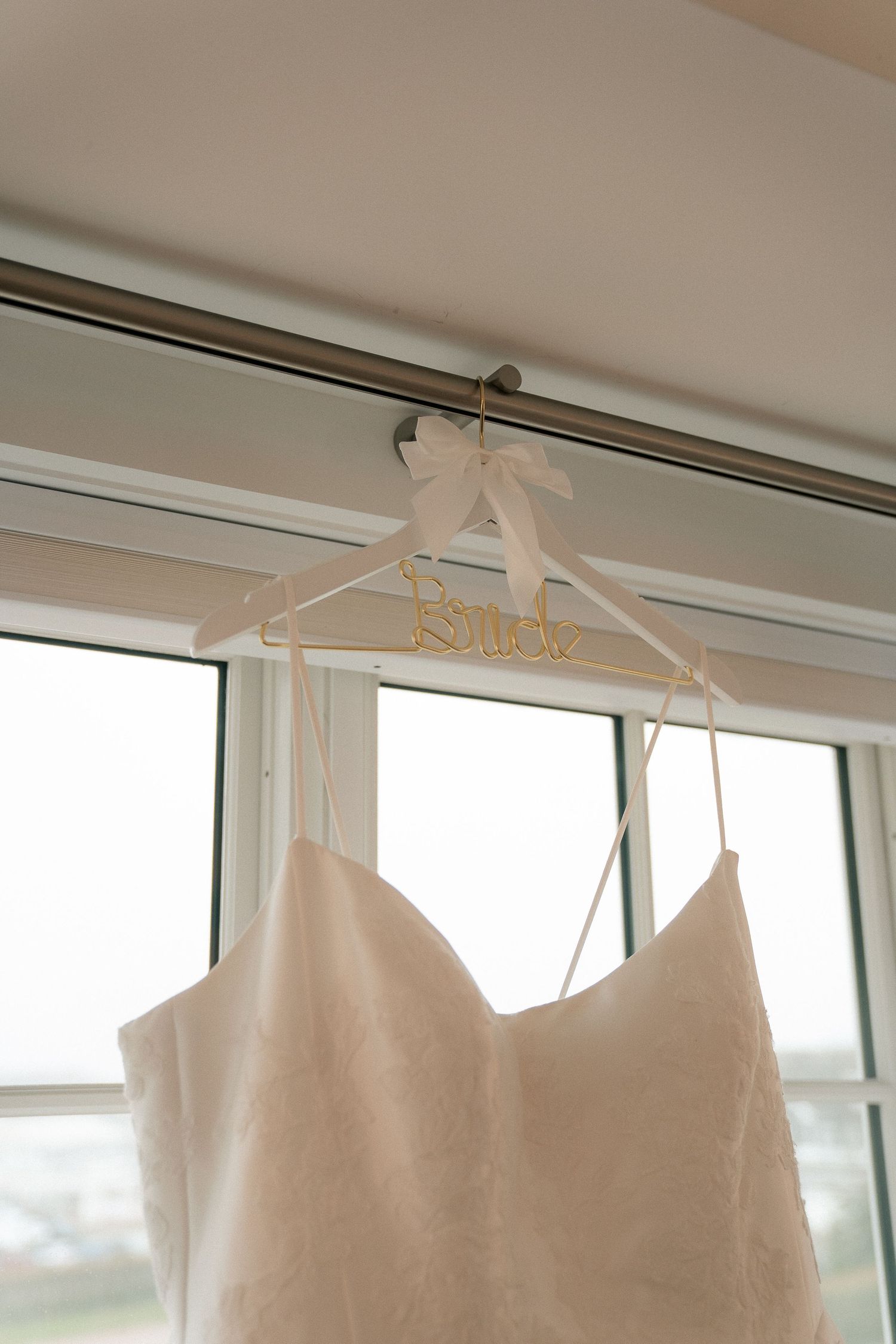 White wedding dress hanging on a custom Bride hanger with gold lettering near a bright window.