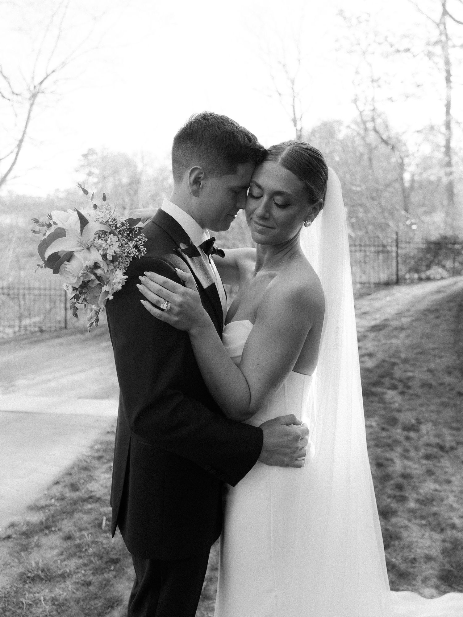 Black and white photo of bride and groom in intimate embrace holding white wedding bouquet.