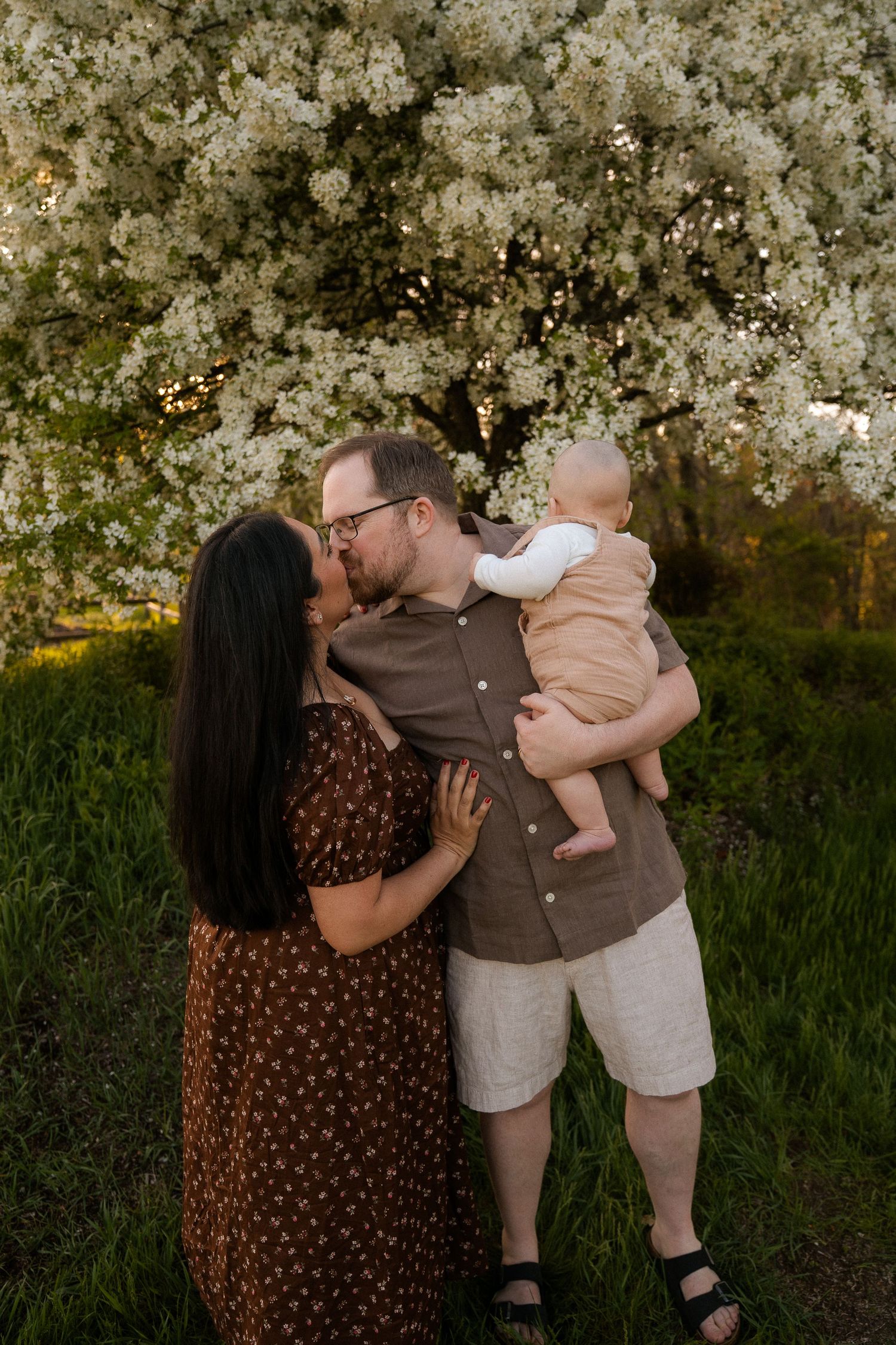 A couple kisses while holding their baby in front of a blooming white flowering tree outdoors.