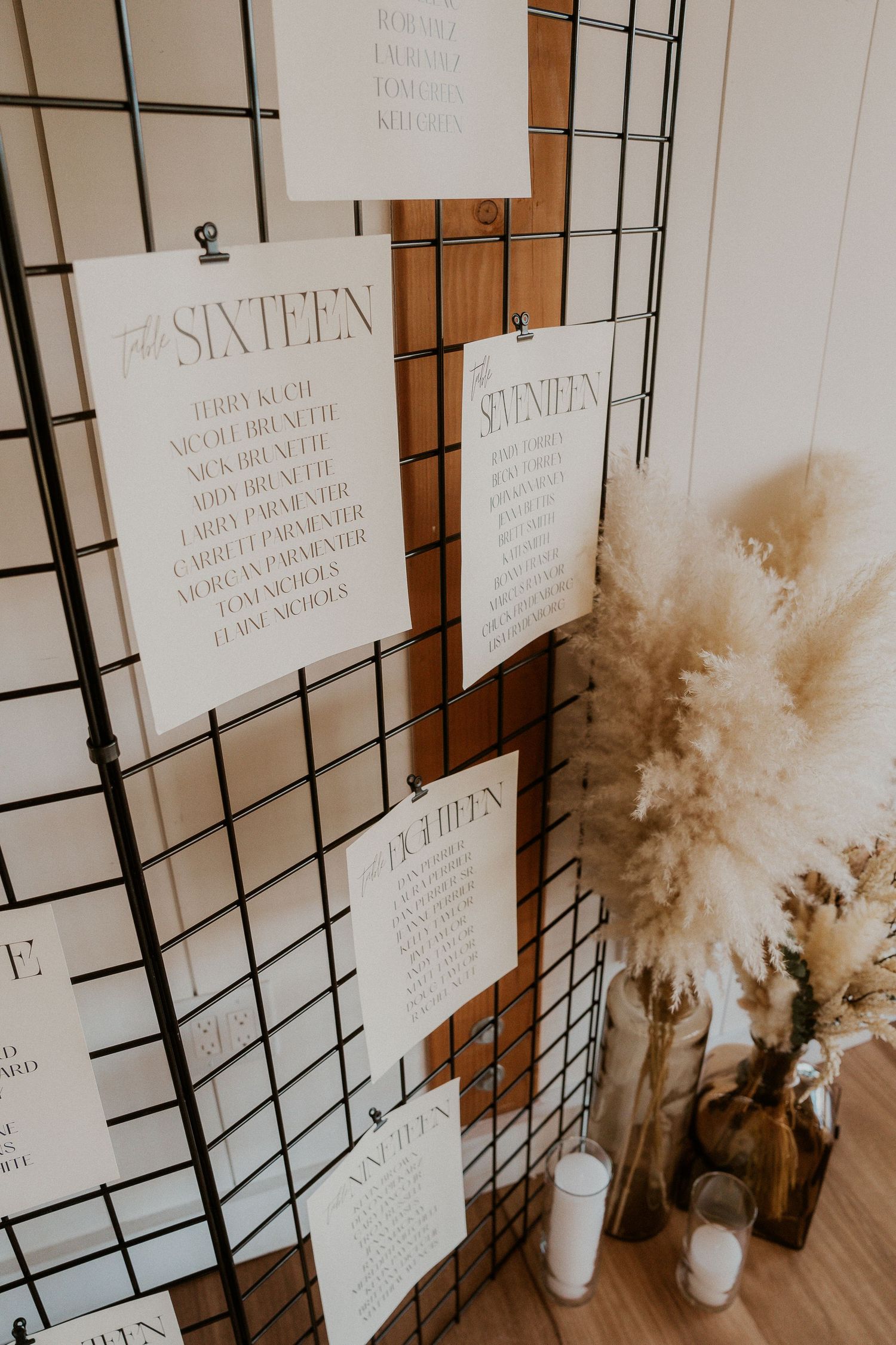 Wedding seating chart display with white paper cards arranged on a grid wall, accented by dried pampas grass arrangements.