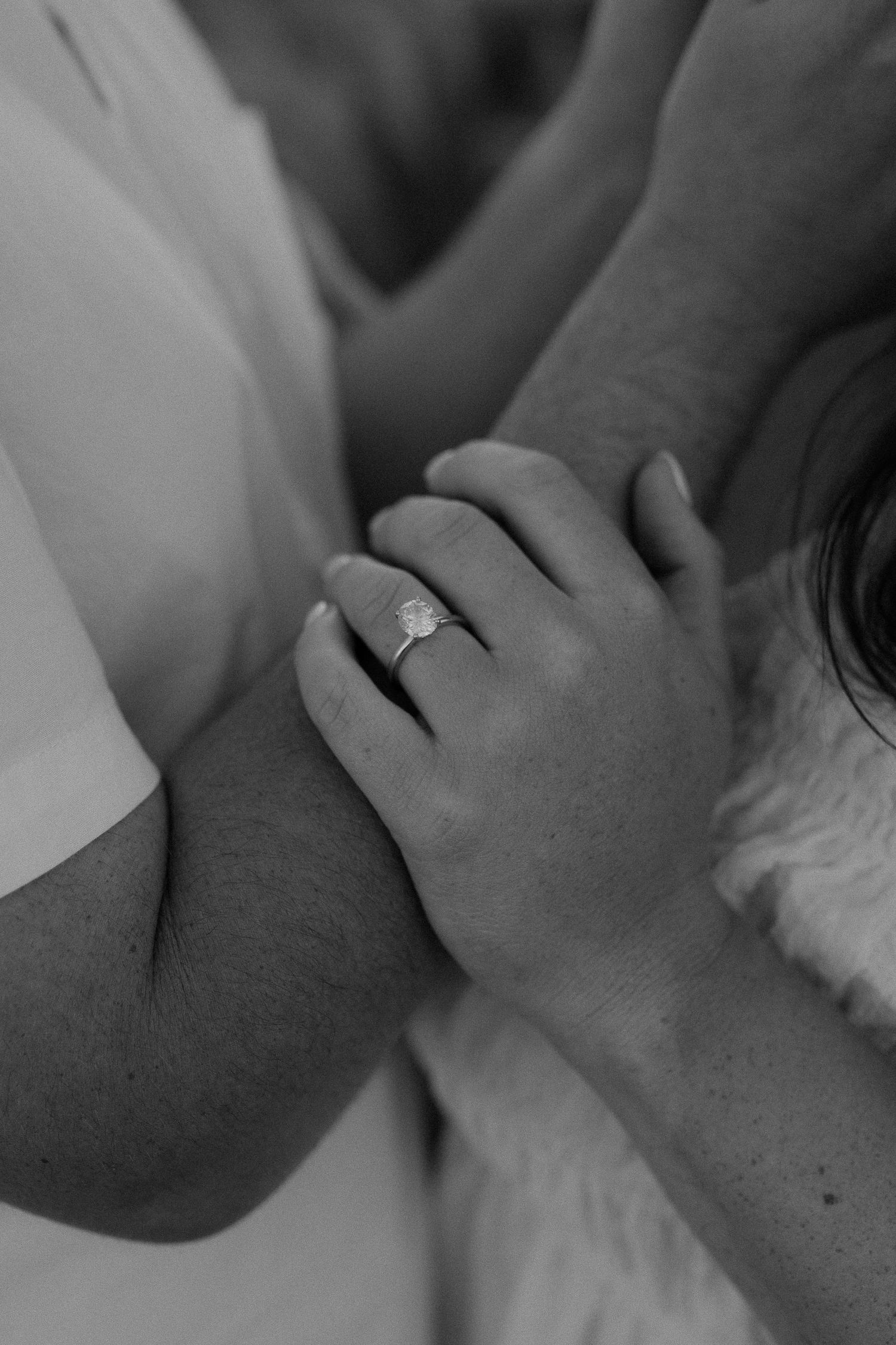 A close-up black and white photo showing hands with an engagement ring in soft focus.