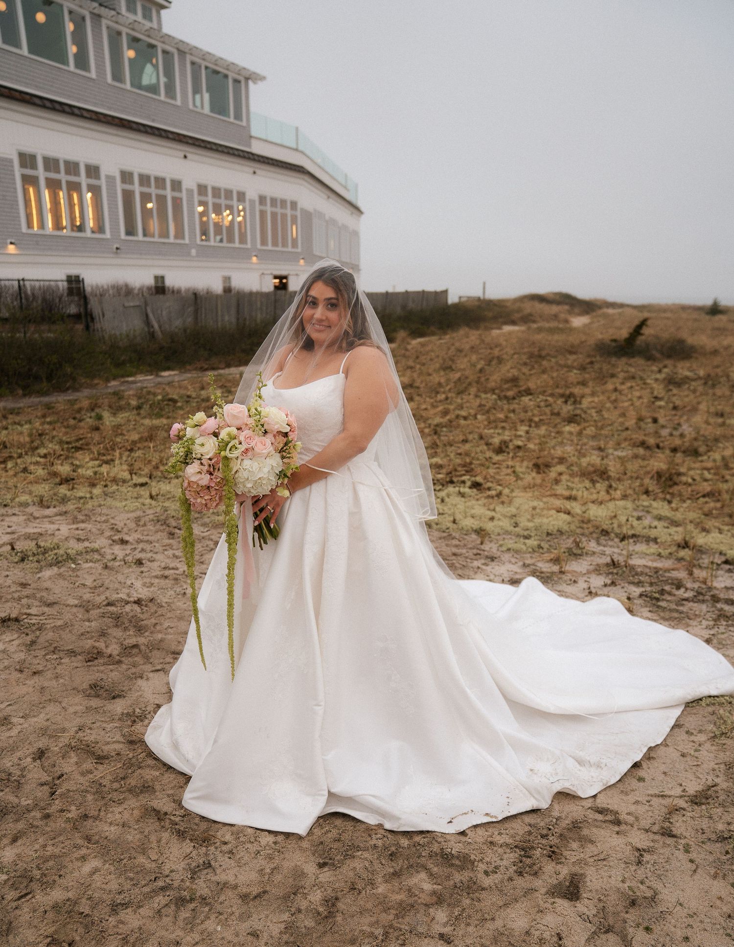 Bride in white ball gown with veil holds pink bouquet on beach near coastal venue.