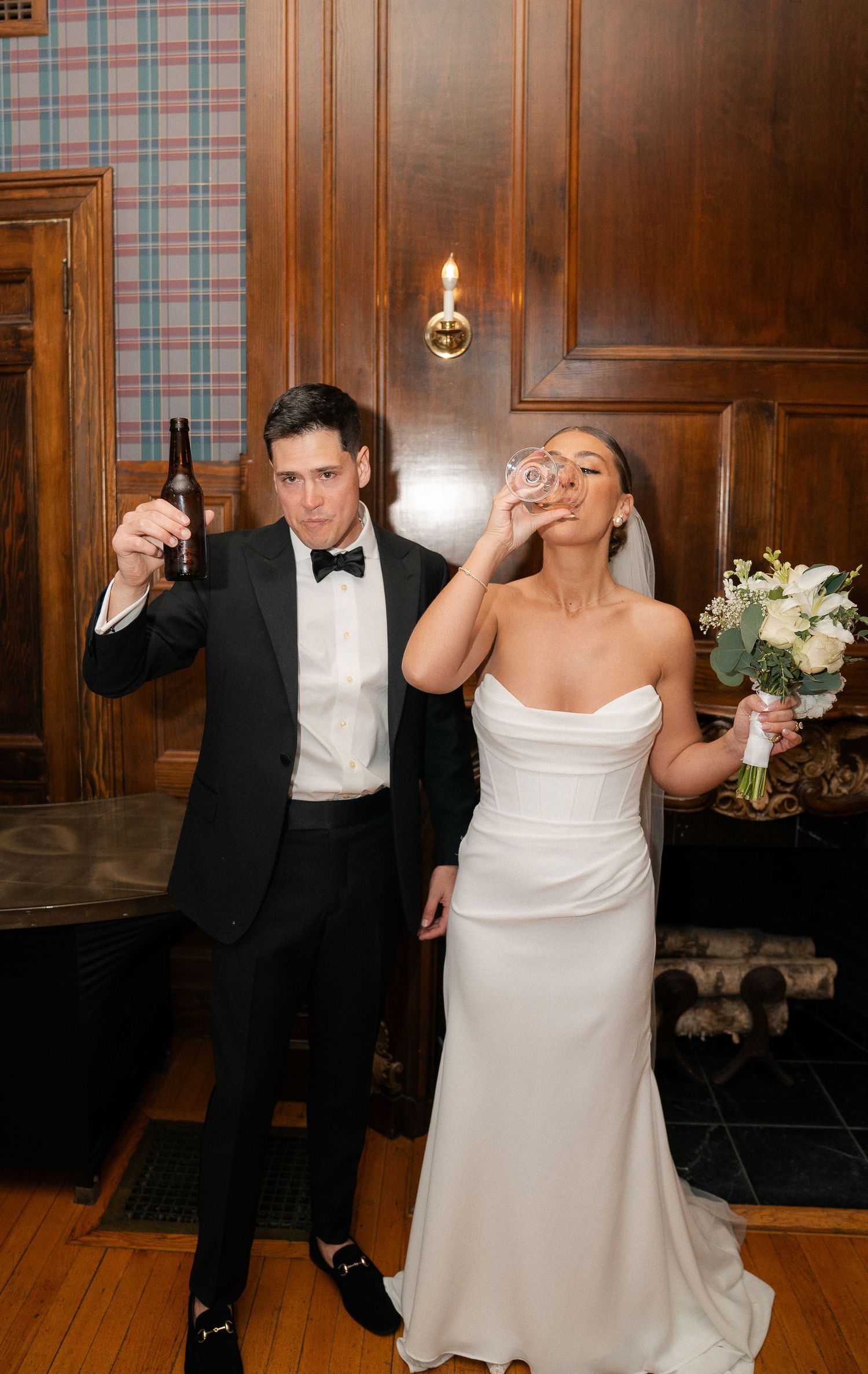 Bride in strapless white gown drinks from a glass while groom in black tuxedo raises a beer bottle in celebration.