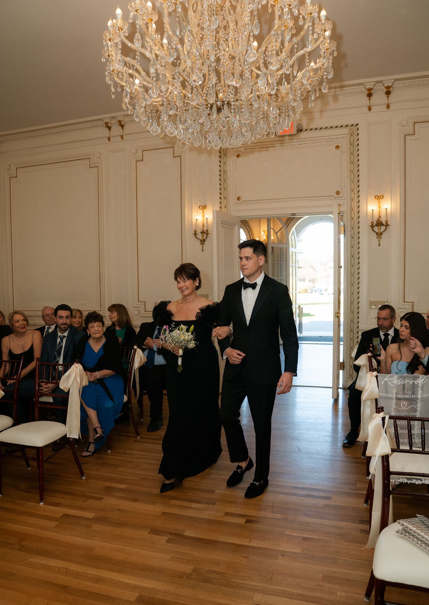 Elegant wedding ceremony with couple walking down aisle under crystal chandelier in a grand white and gold ballroom.