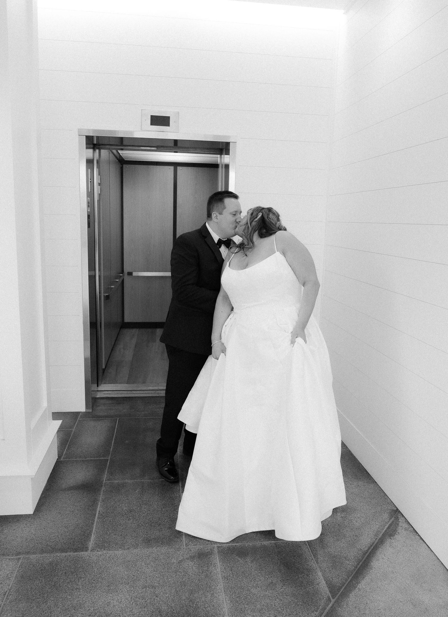 Black and white photo of a bride and groom sharing a kiss in front of an elevator on their wedding day.