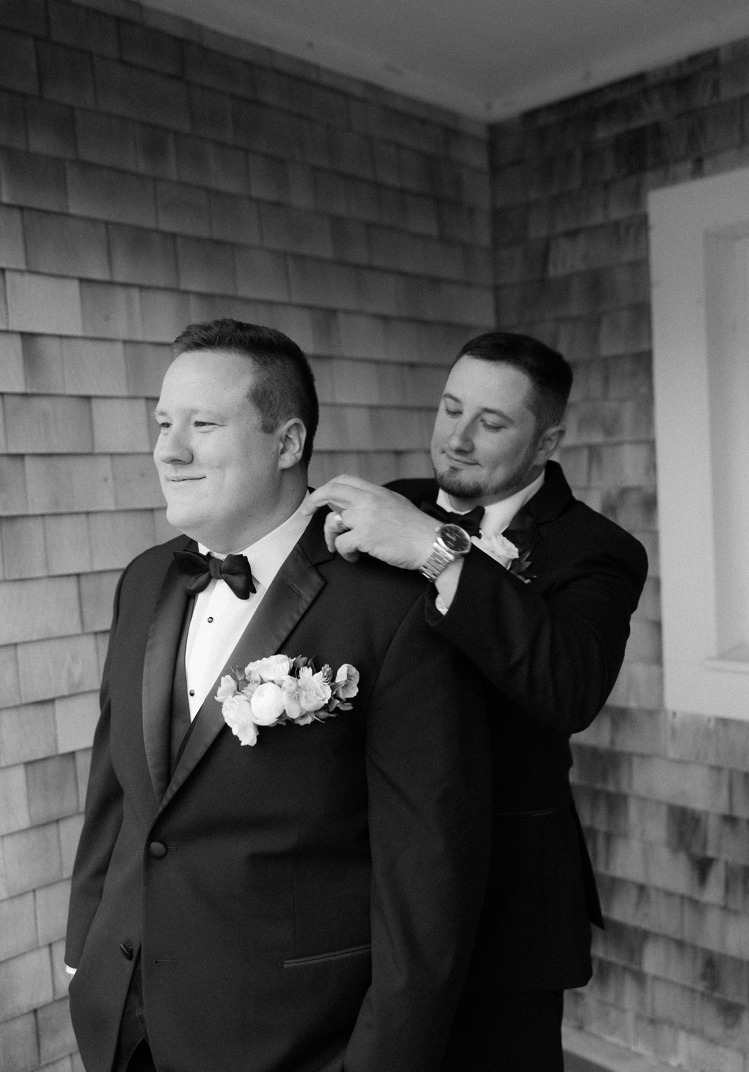 Black and white photo of groom in tuxedo being helped by groomsman outside shingled building.