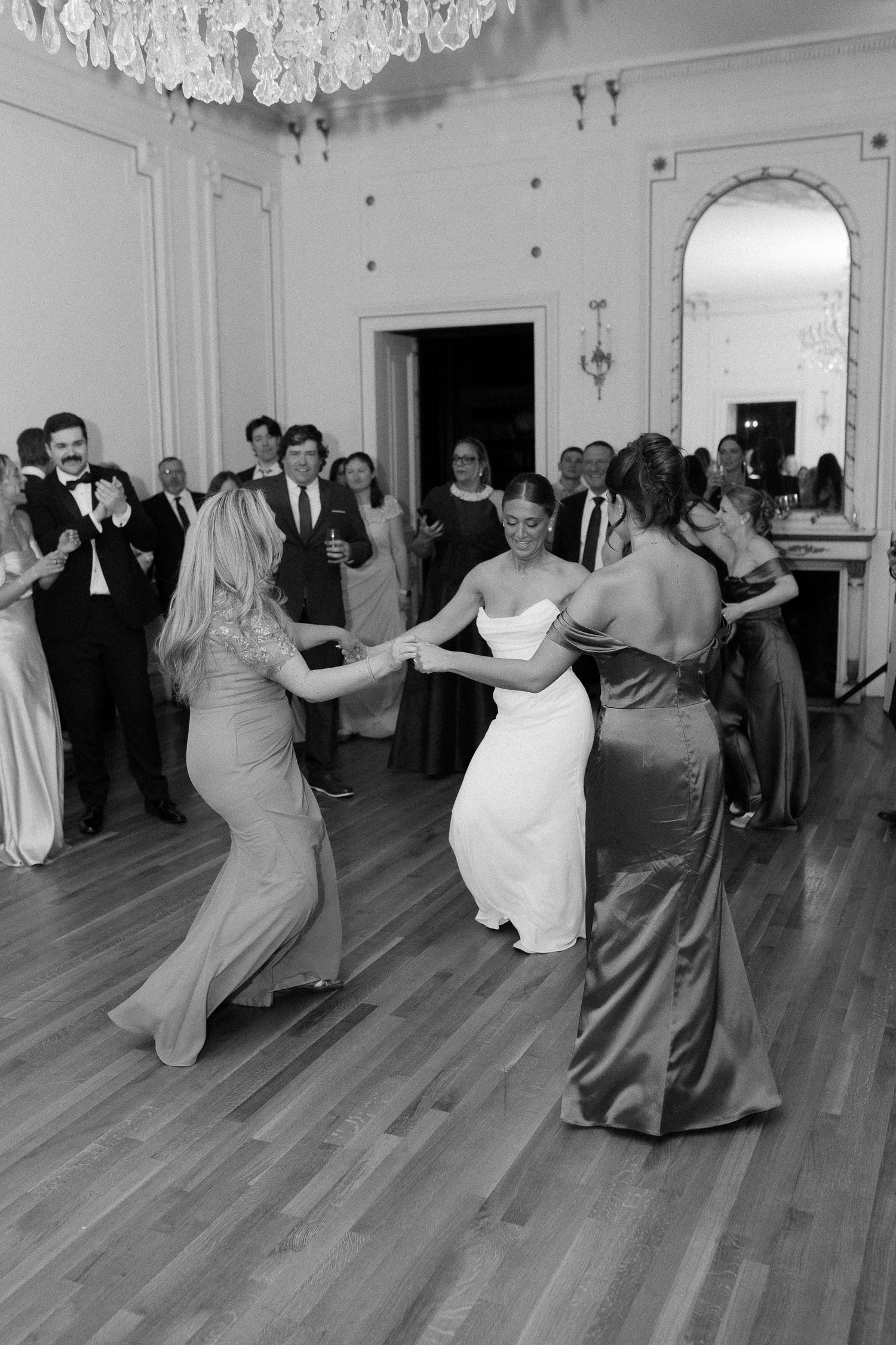 Bride dancing with bridesmaids in elegant gowns on wooden dance floor at wedding reception, guests watching in background.