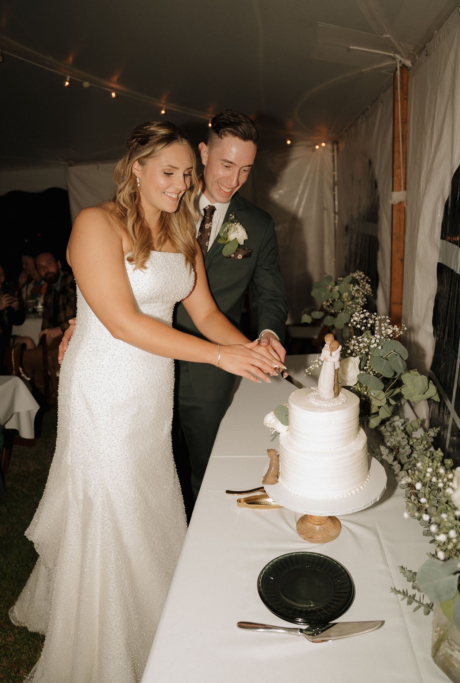 A couple cuts their elegant white wedding cake decorated with greenery.