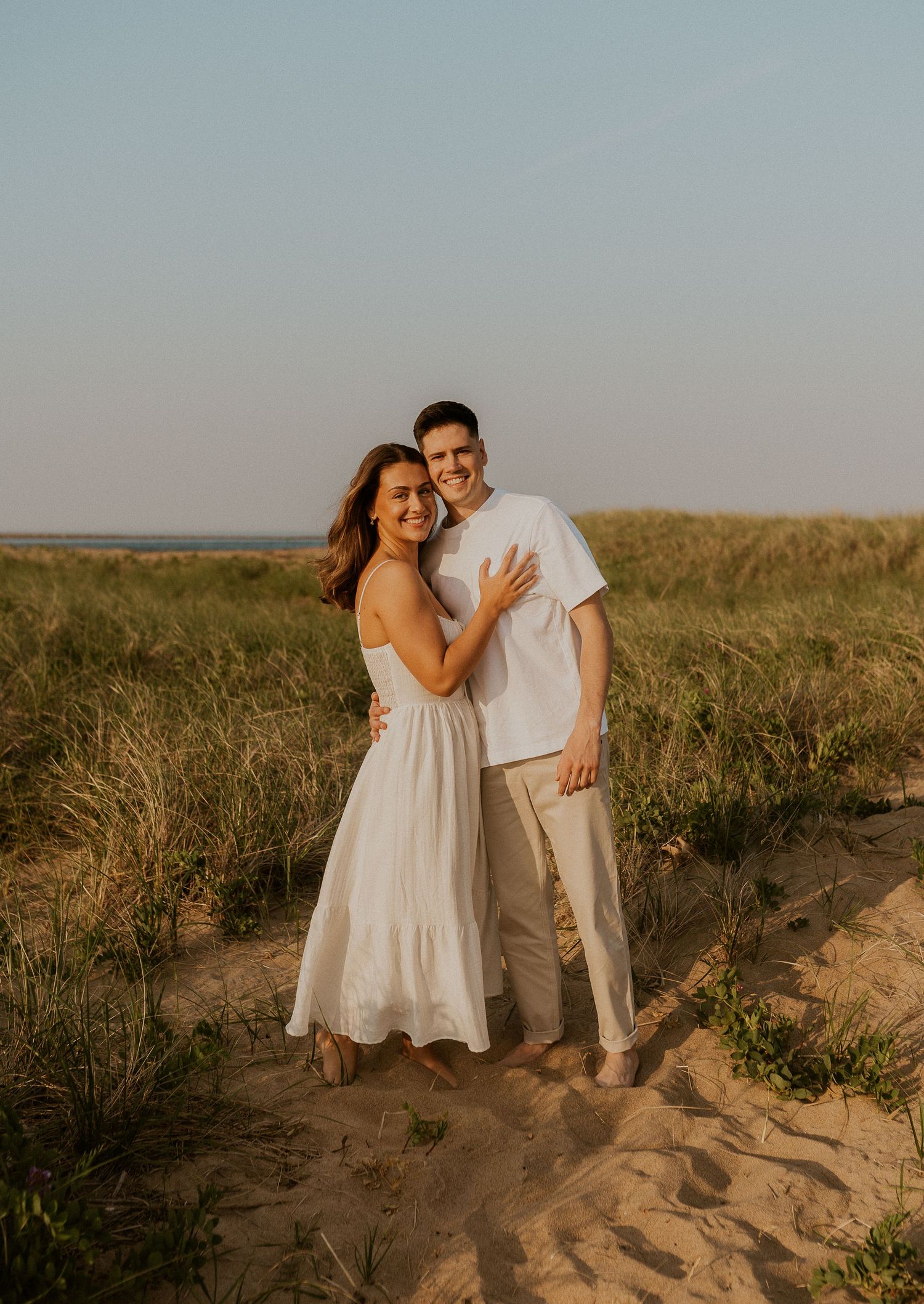 A romantic couple in white outfits embraces on sandy dunes during golden hour with tall grass in the background.