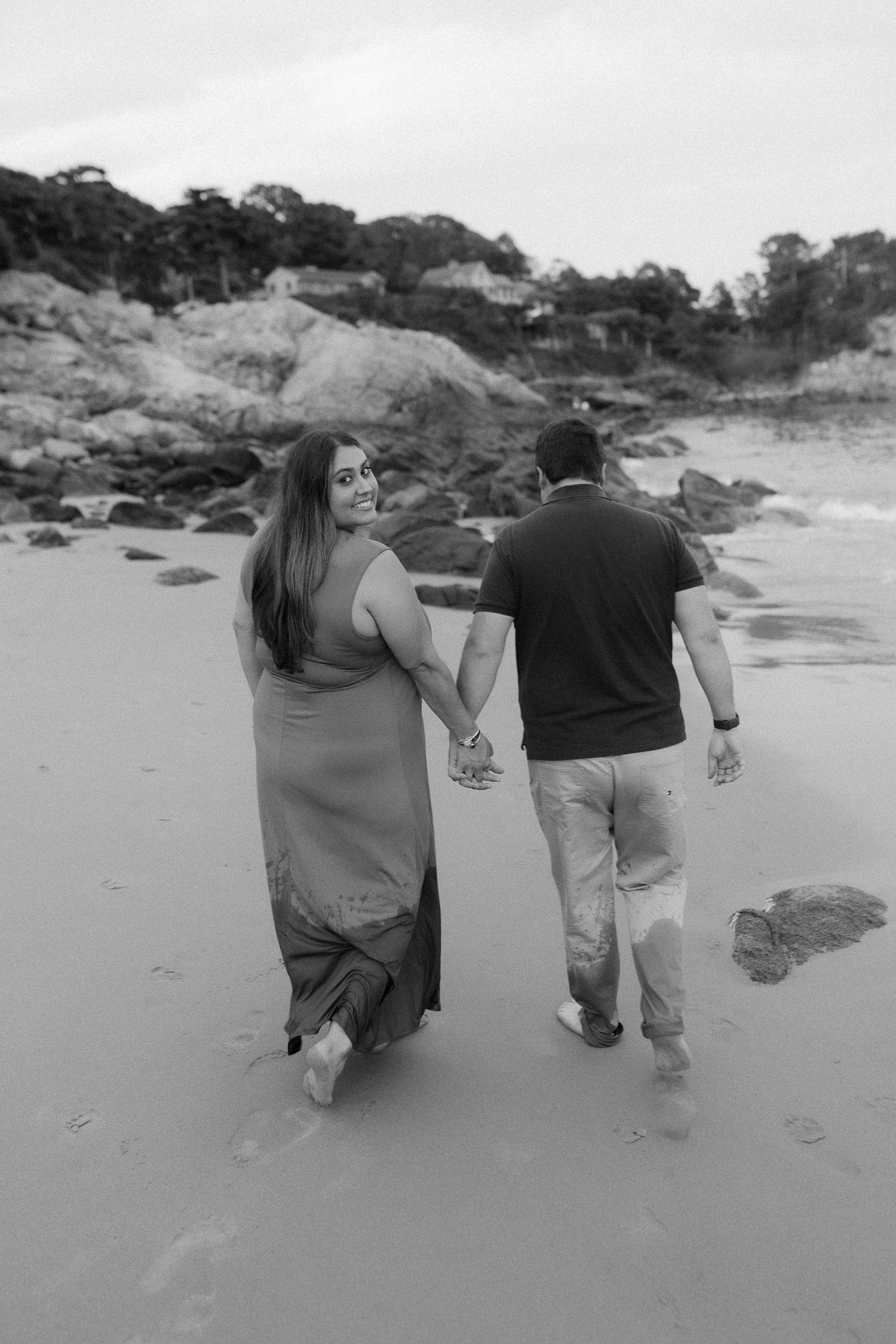 Couple walking hand in hand along a sandy beach in black and white.