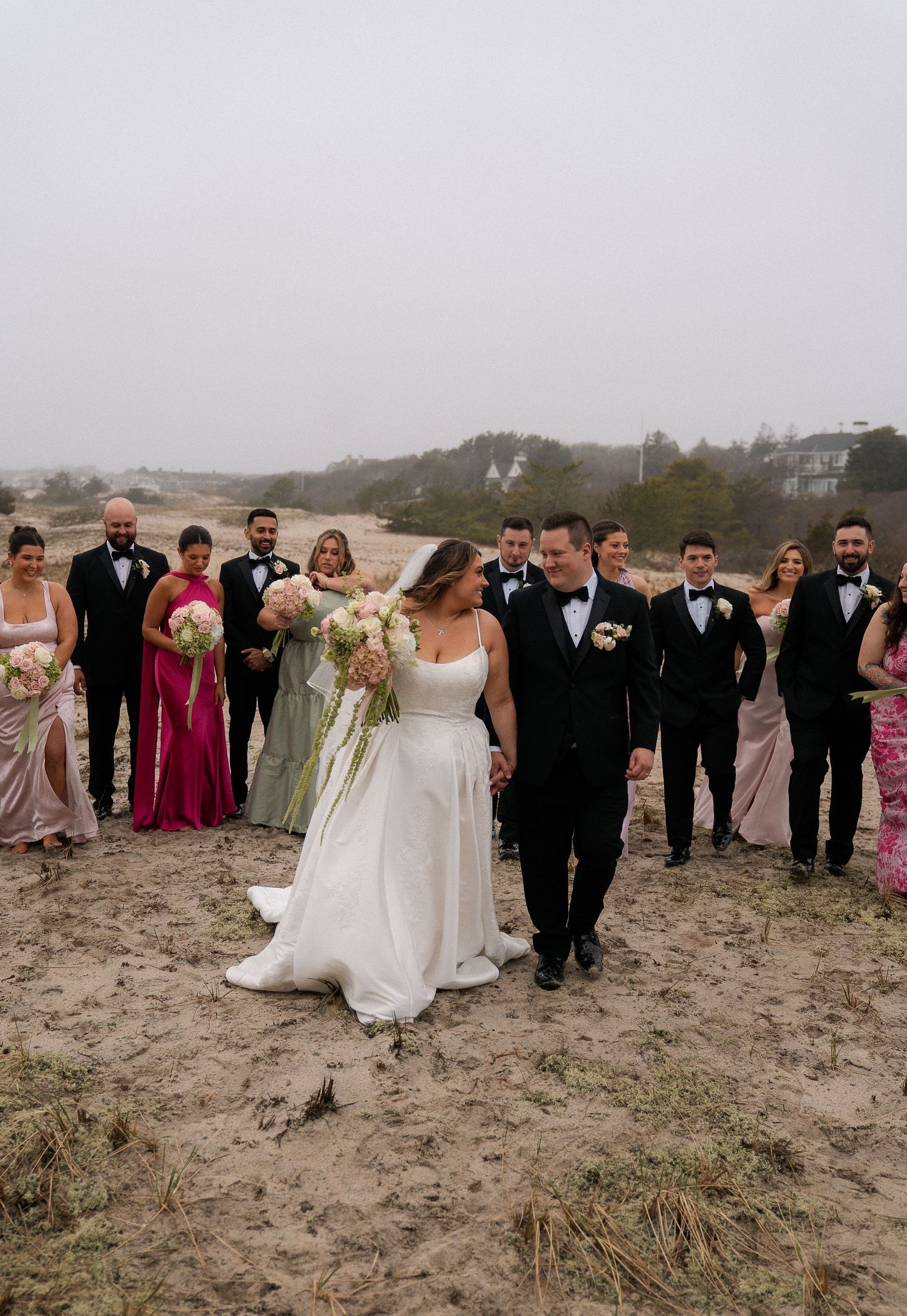 Wedding party posing on sandy beach with bride and groom in center on overcast day.