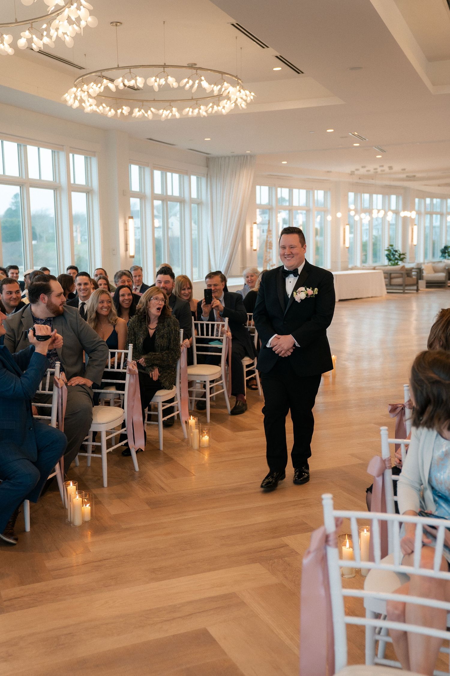 Groom in black tuxedo walks down aisle at elegant indoor wedding ceremony with chandeliers and white chiavari chairs.