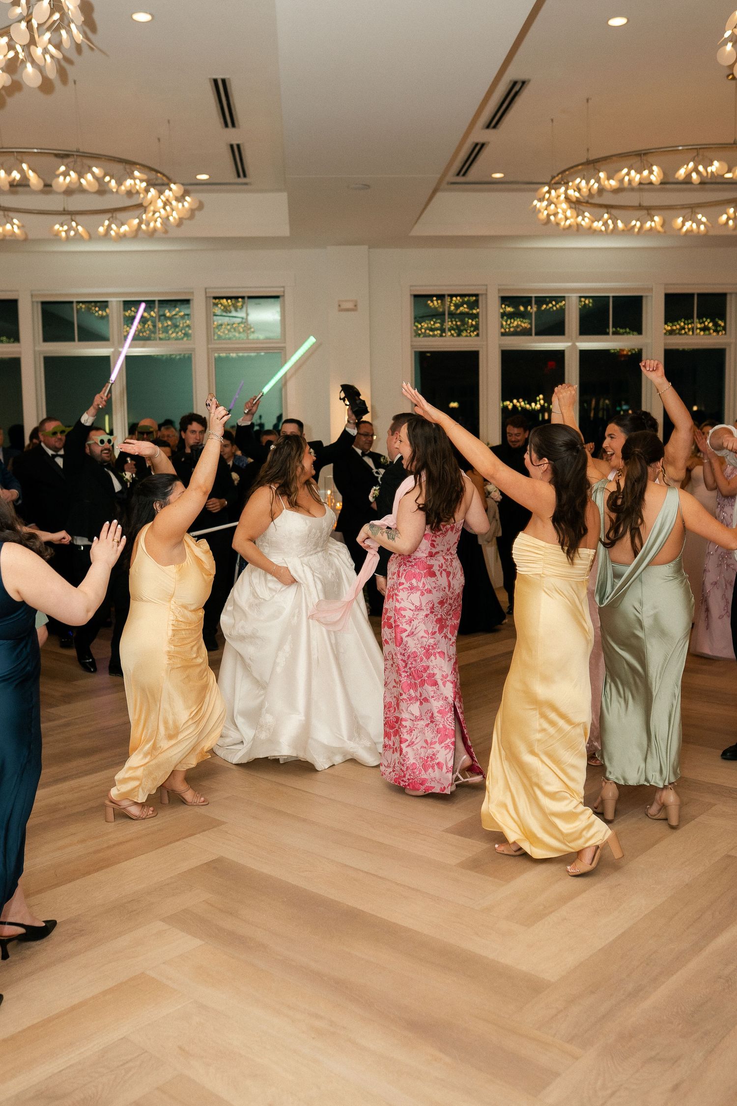 Bride and bridesmaids celebrate on dance floor with light-up sabers at lively wedding reception.