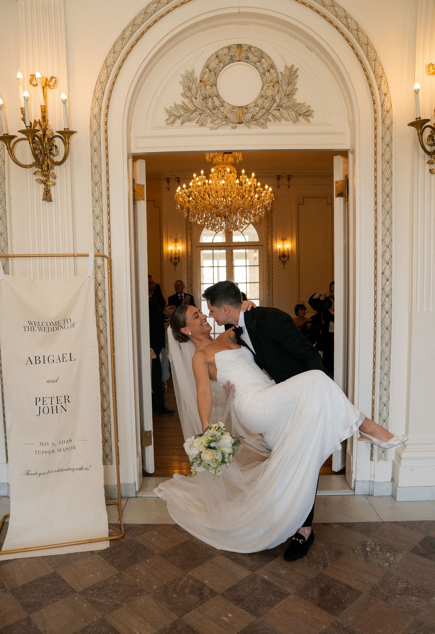 Groom lifts bride for a romantic kiss in an ornate doorway beside Abigael and Peter John wedding sign.